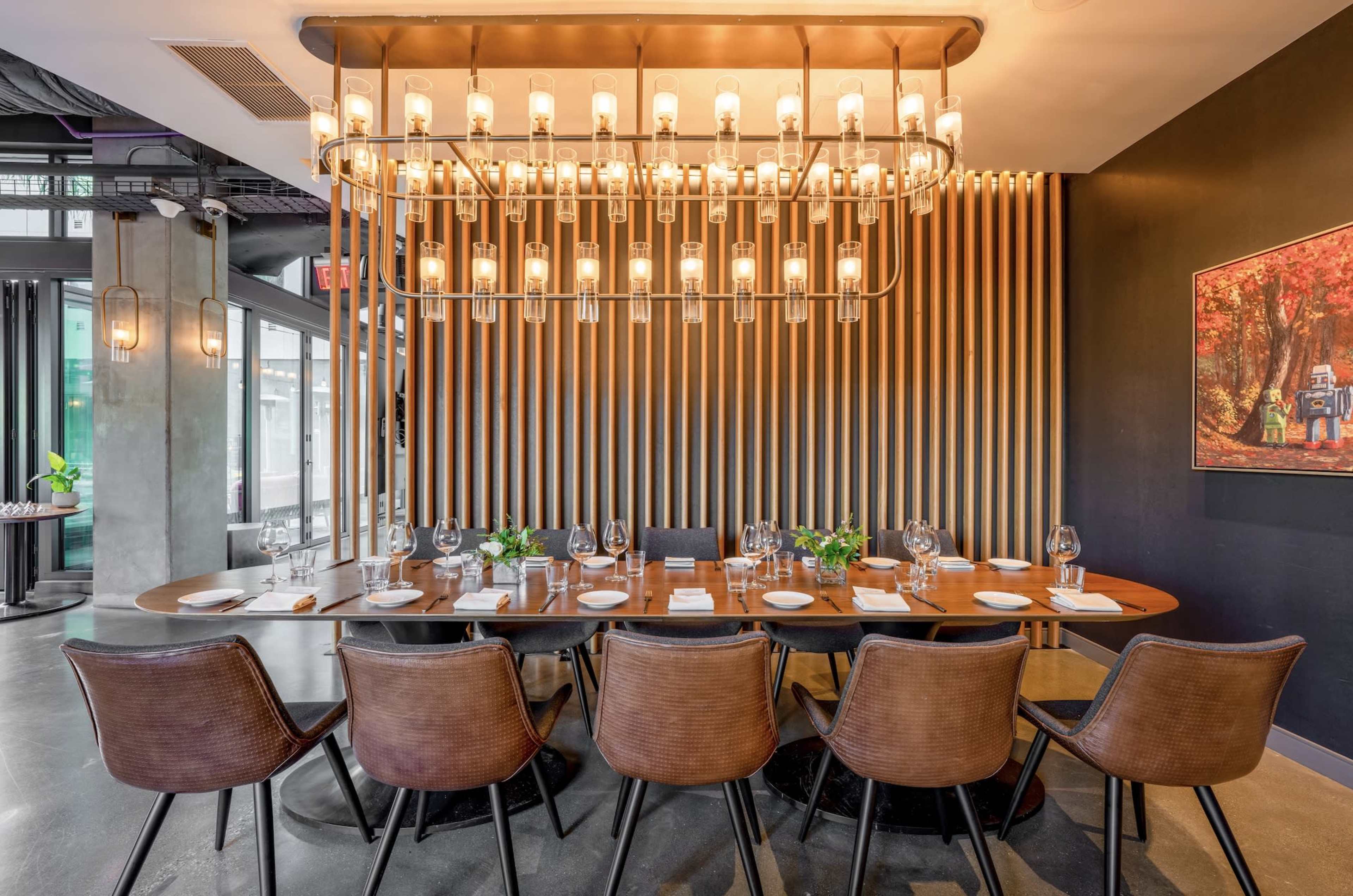 A long wooden table is set for dining with elegant tableware and surrounded by eight chairs, beneath a striking chandelier and against a wall featuring wooden slats.