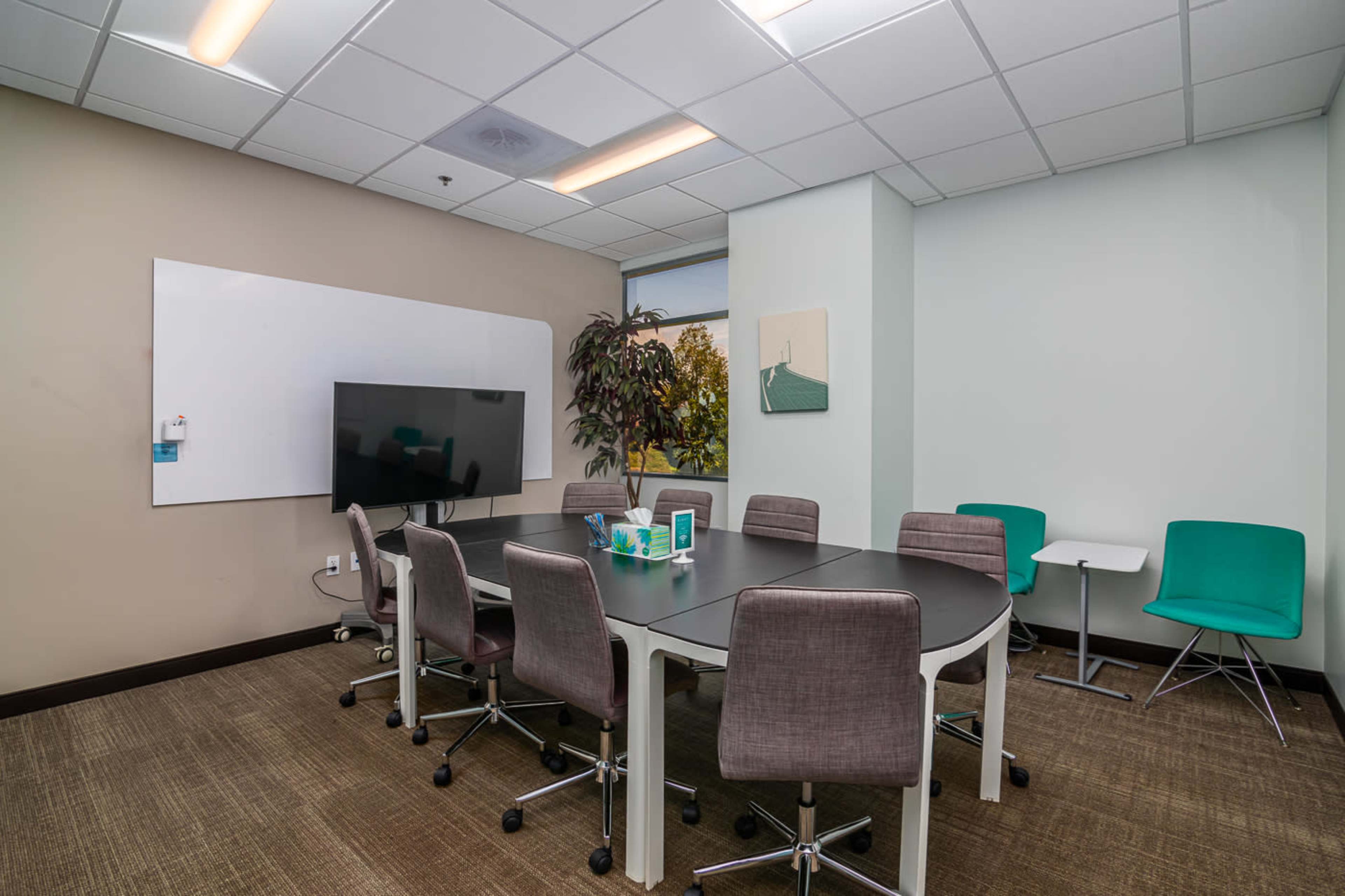 A conference room features a round table surrounded by chairs, a large screen, and a whiteboard on the wall.