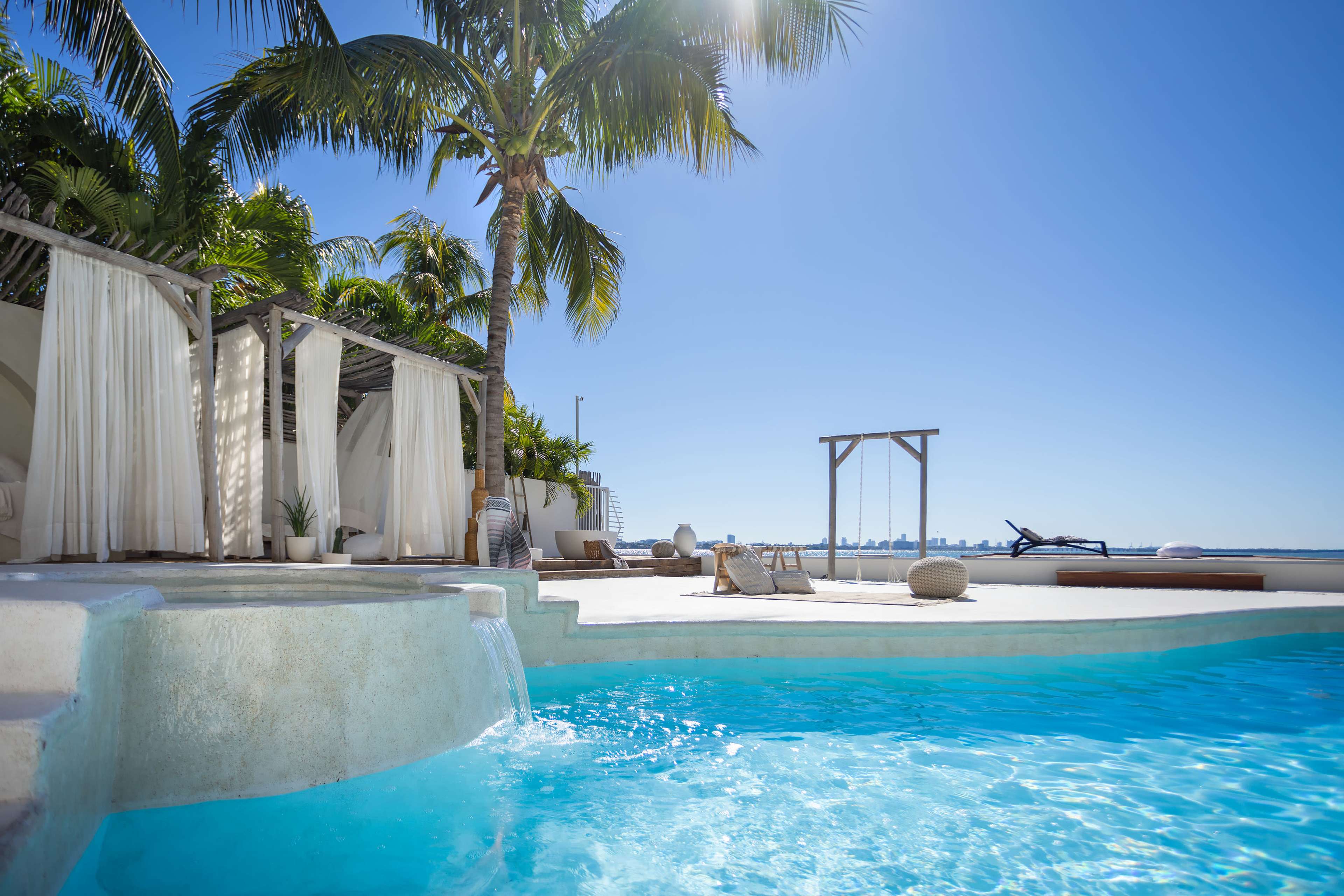 A sparkling blue pool with a waterfall is situated next to a sunlit deck featuring palm trees and lounge chairs.