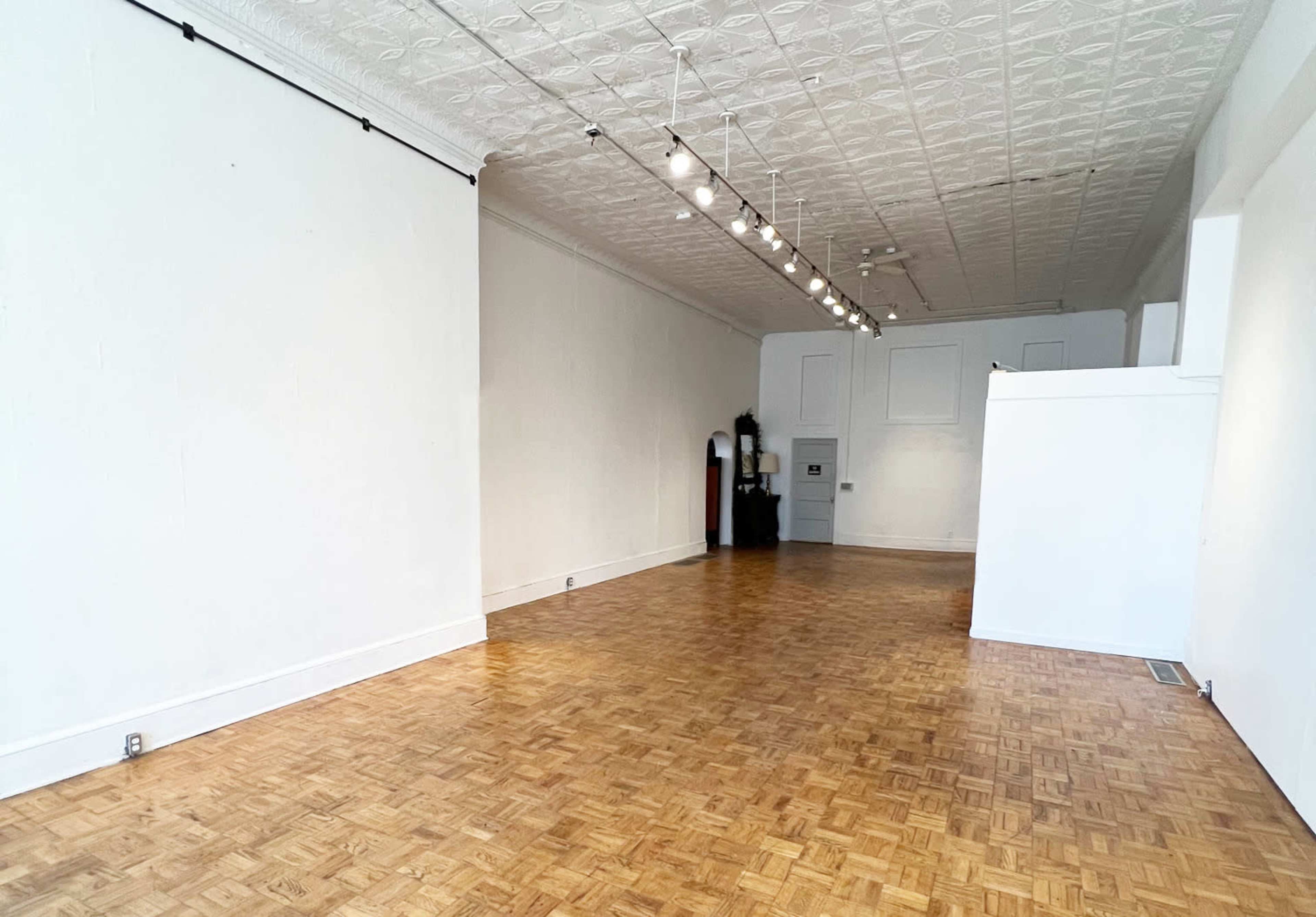 The image shows a spacious, empty room with hardwood flooring and white walls, featuring track lighting along the ceiling.