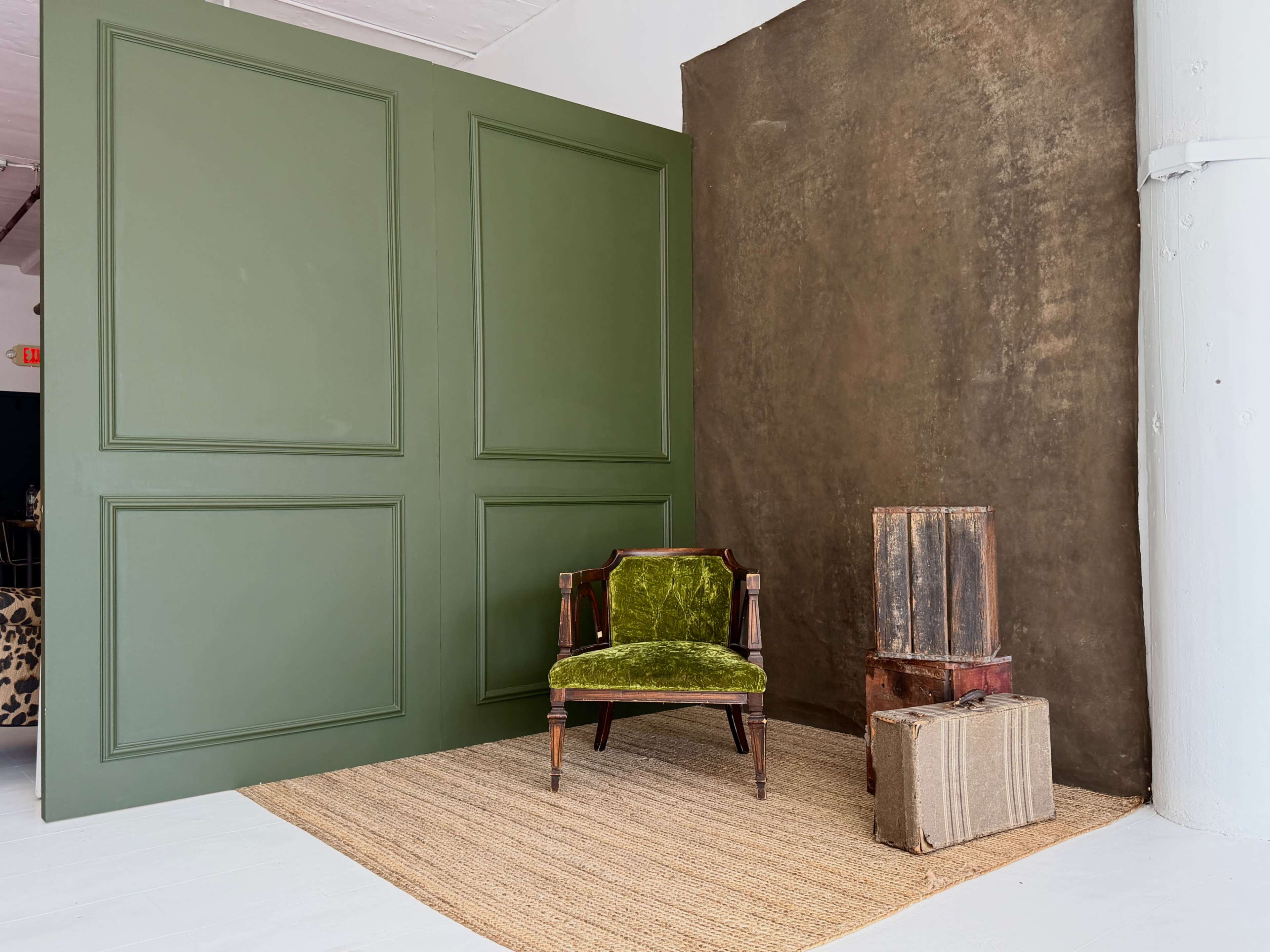 A green-paneled wall serves as a backdrop for a vintage green velvet chair next to stacked wooden crates on a natural fiber rug.
