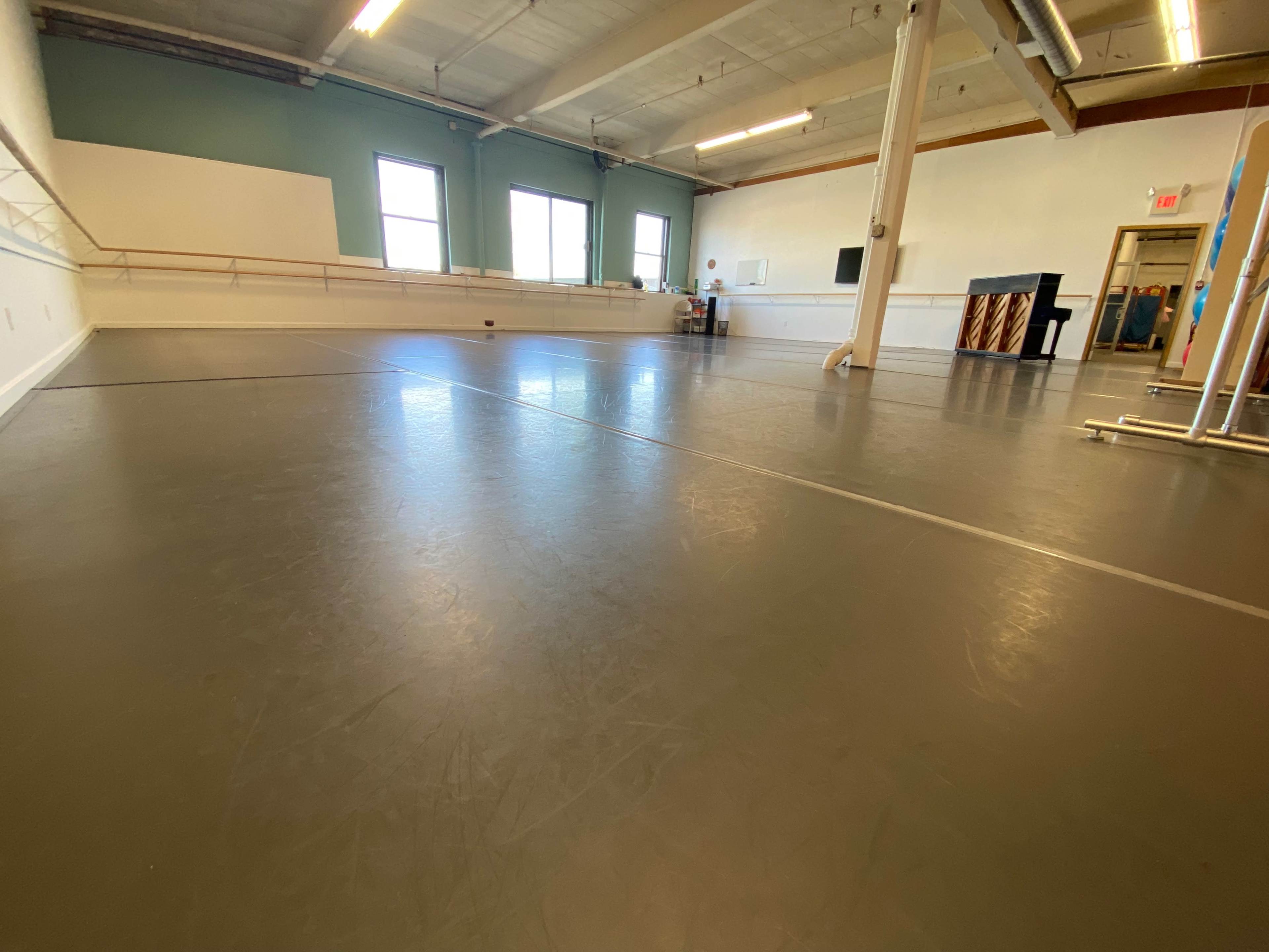 A spacious dance studio with a smooth gray floor, mirrored walls, and a piano in the corner.