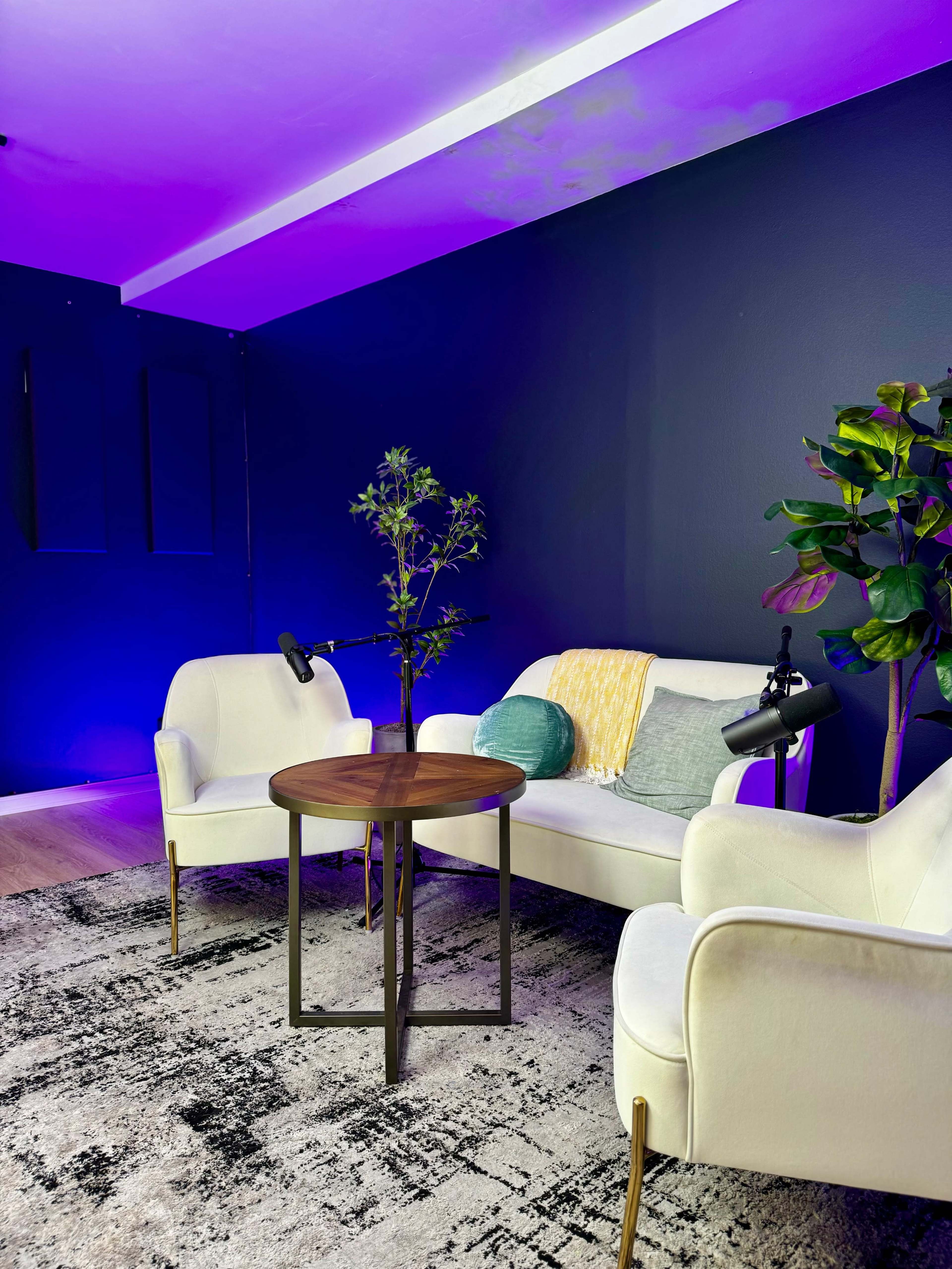 A cozy seating area with two white armchairs, a small round wooden table, and a decorative plant, all set against a dark wall illuminated by purple light.