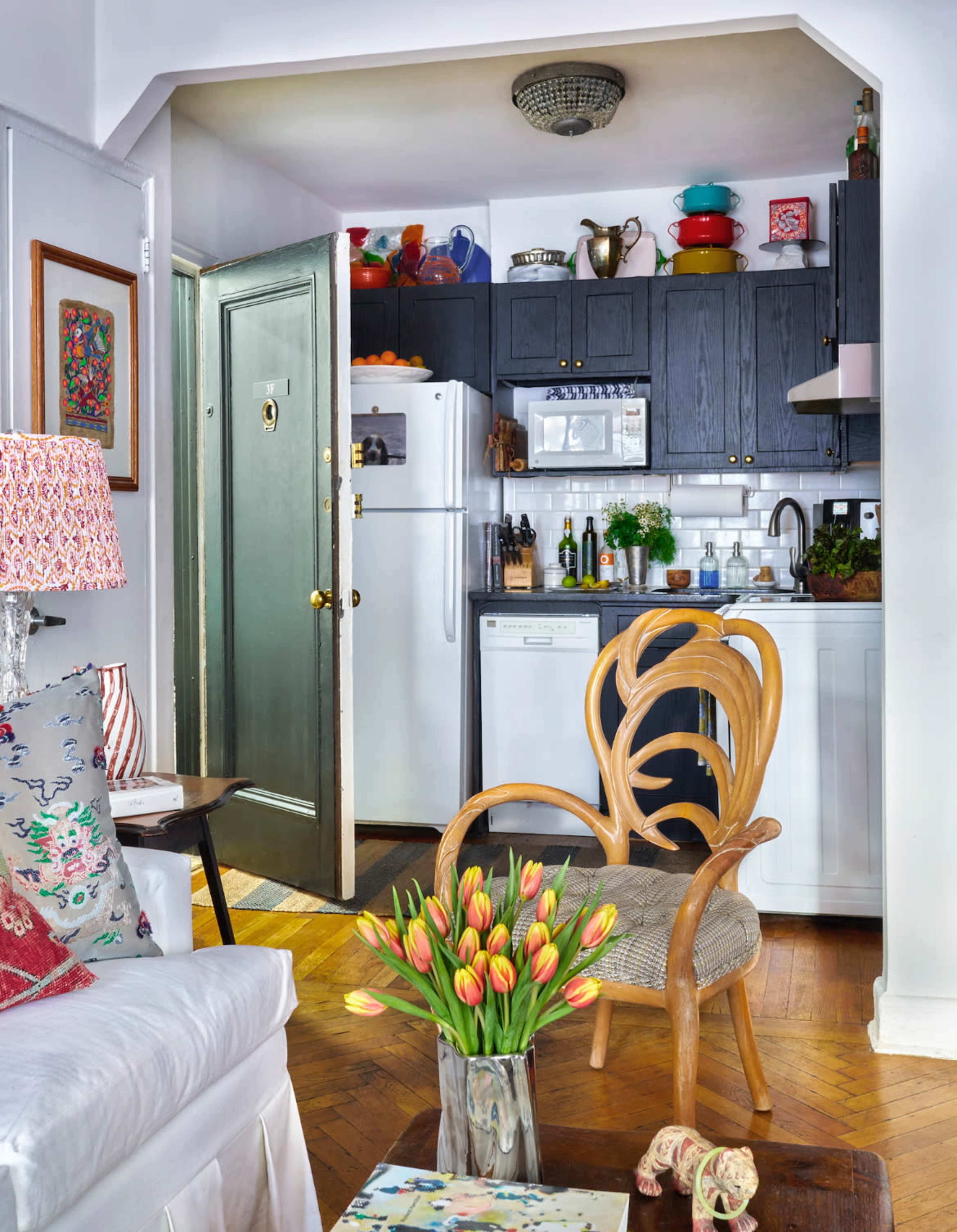 The image shows a well-lit, compact apartment kitchen adjoining a cozy living area furnished with a decorative chair and a vase of tulips.