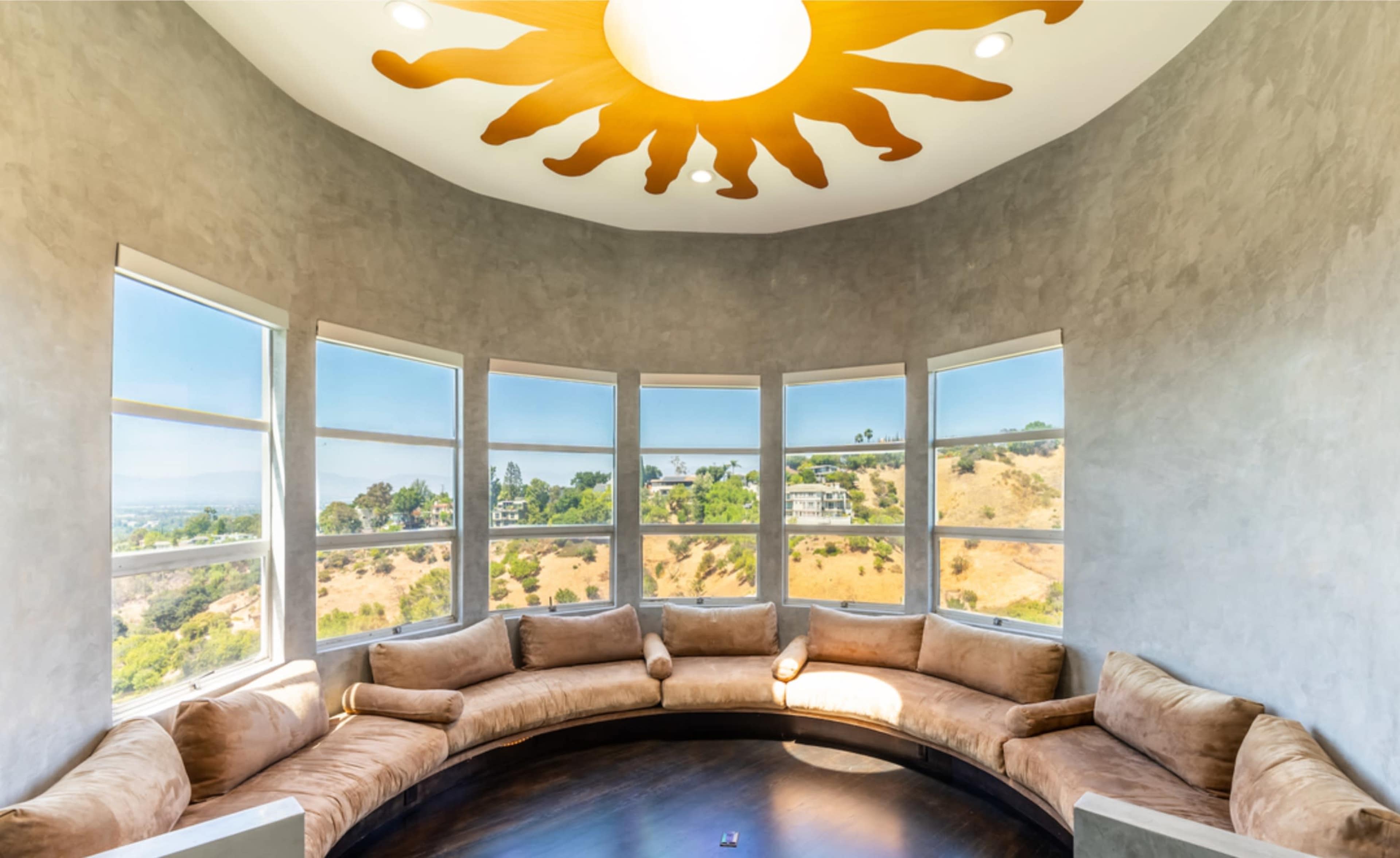 A rounded seating area with a sun-shaped ceiling light, surrounded by large windows that overlook a hilly landscape.