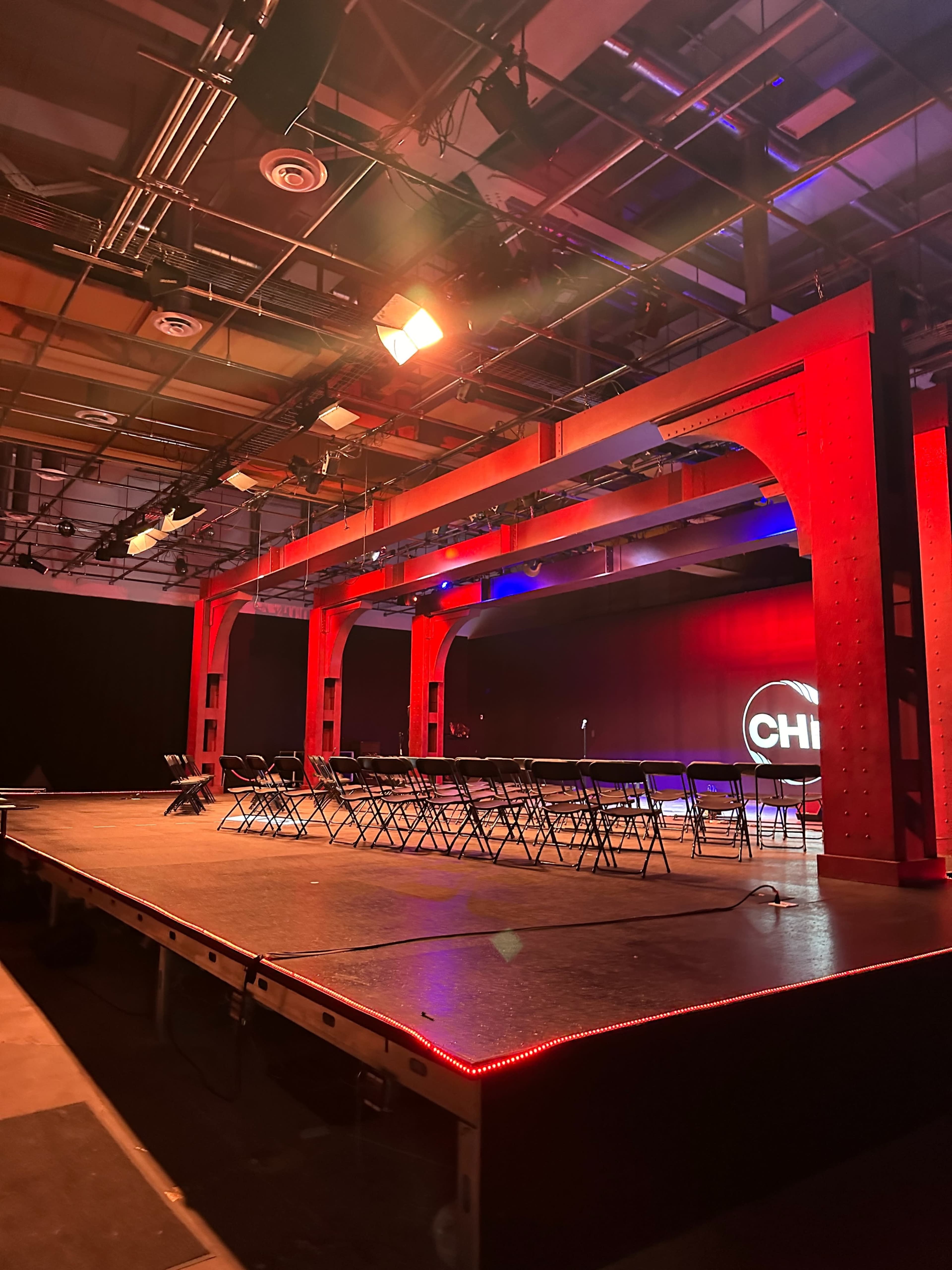 The image shows a stage set up with a row of chairs under red lighting and a circular logo at the back.