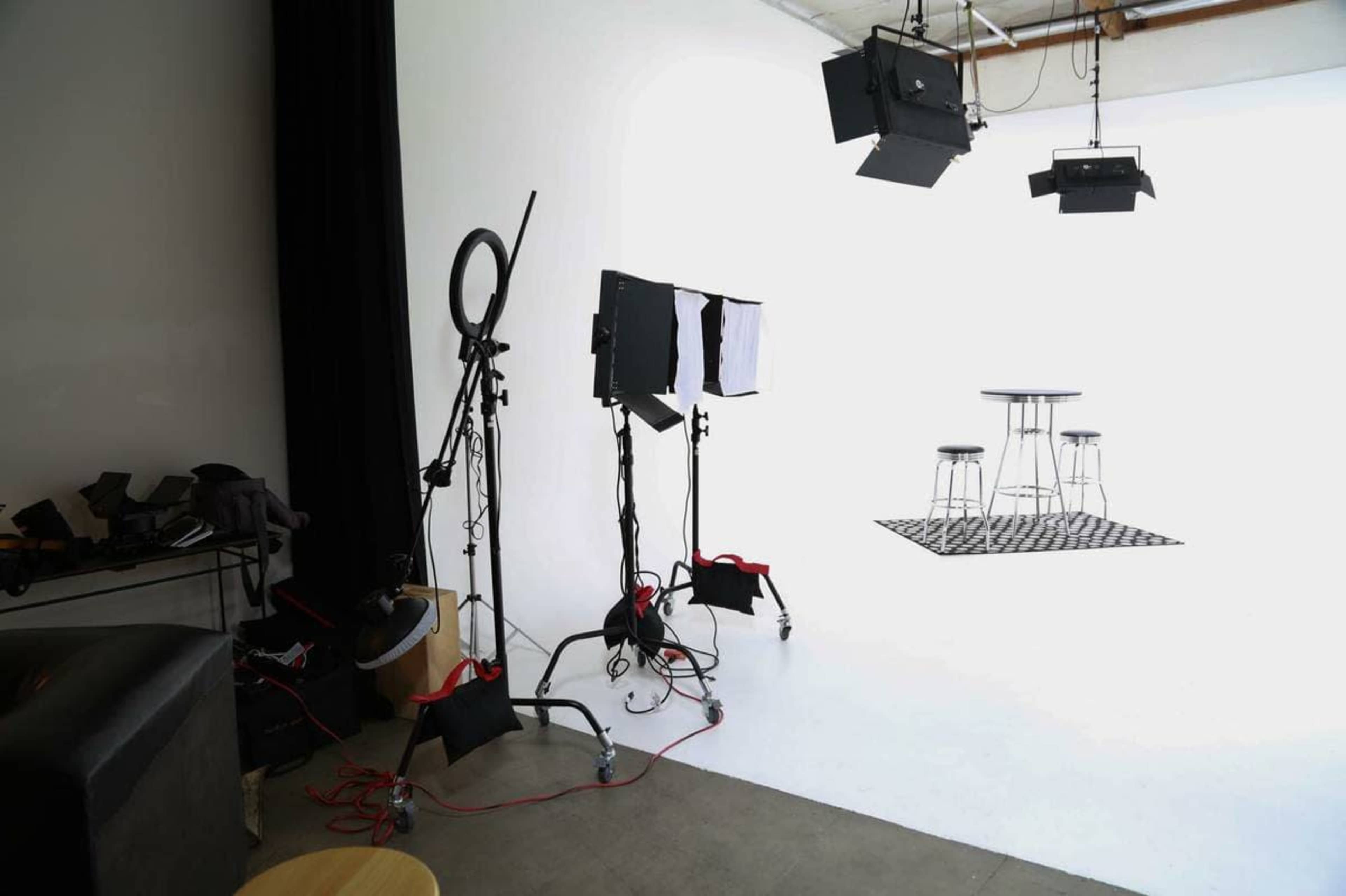 A photography studio is set up with lighting equipment, a checkered rug, and two stools against a white backdrop.