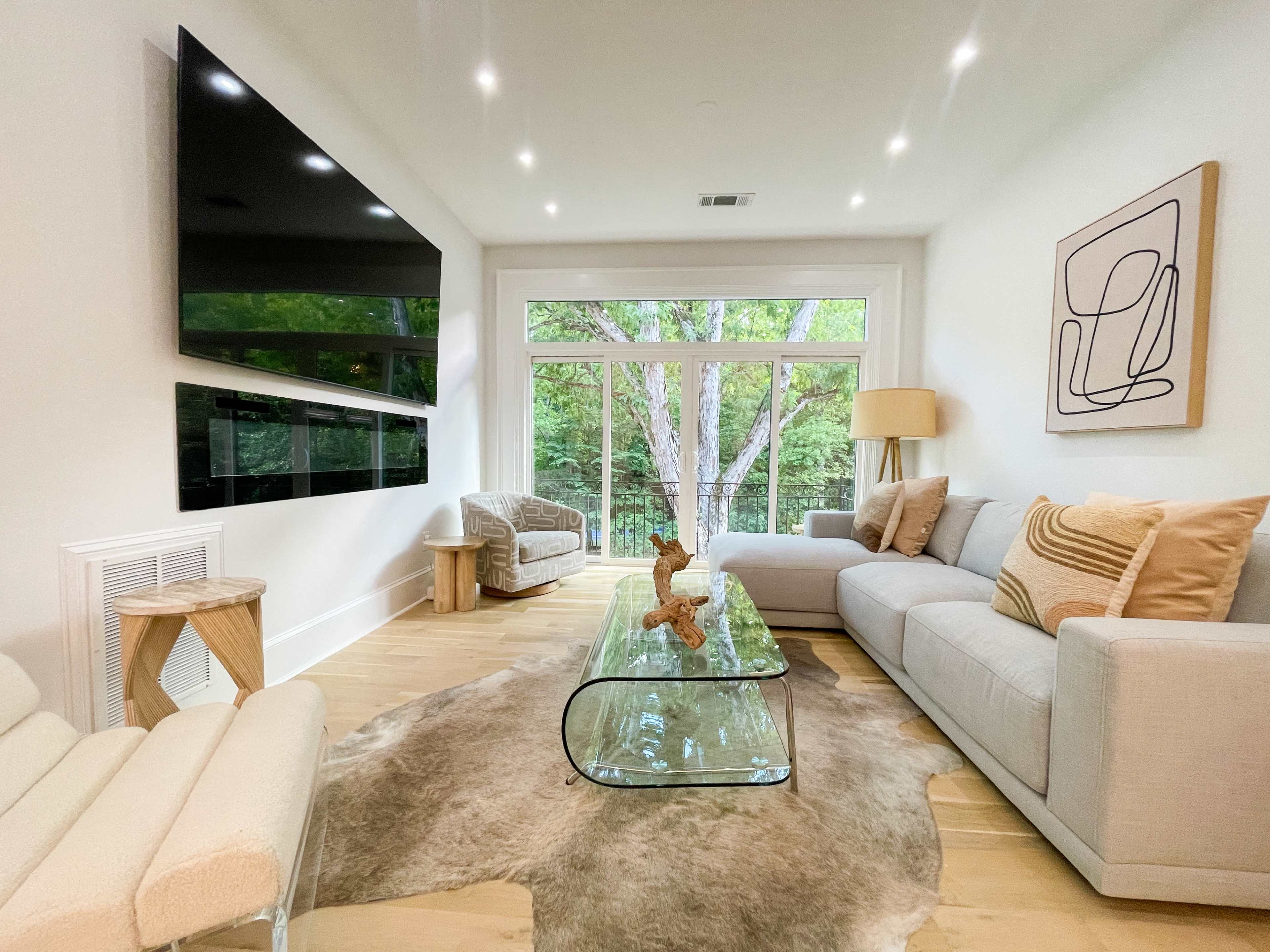 A modern living room features a light-colored couch, a glass coffee table on a rug, and large windows that overlook a wooded area.