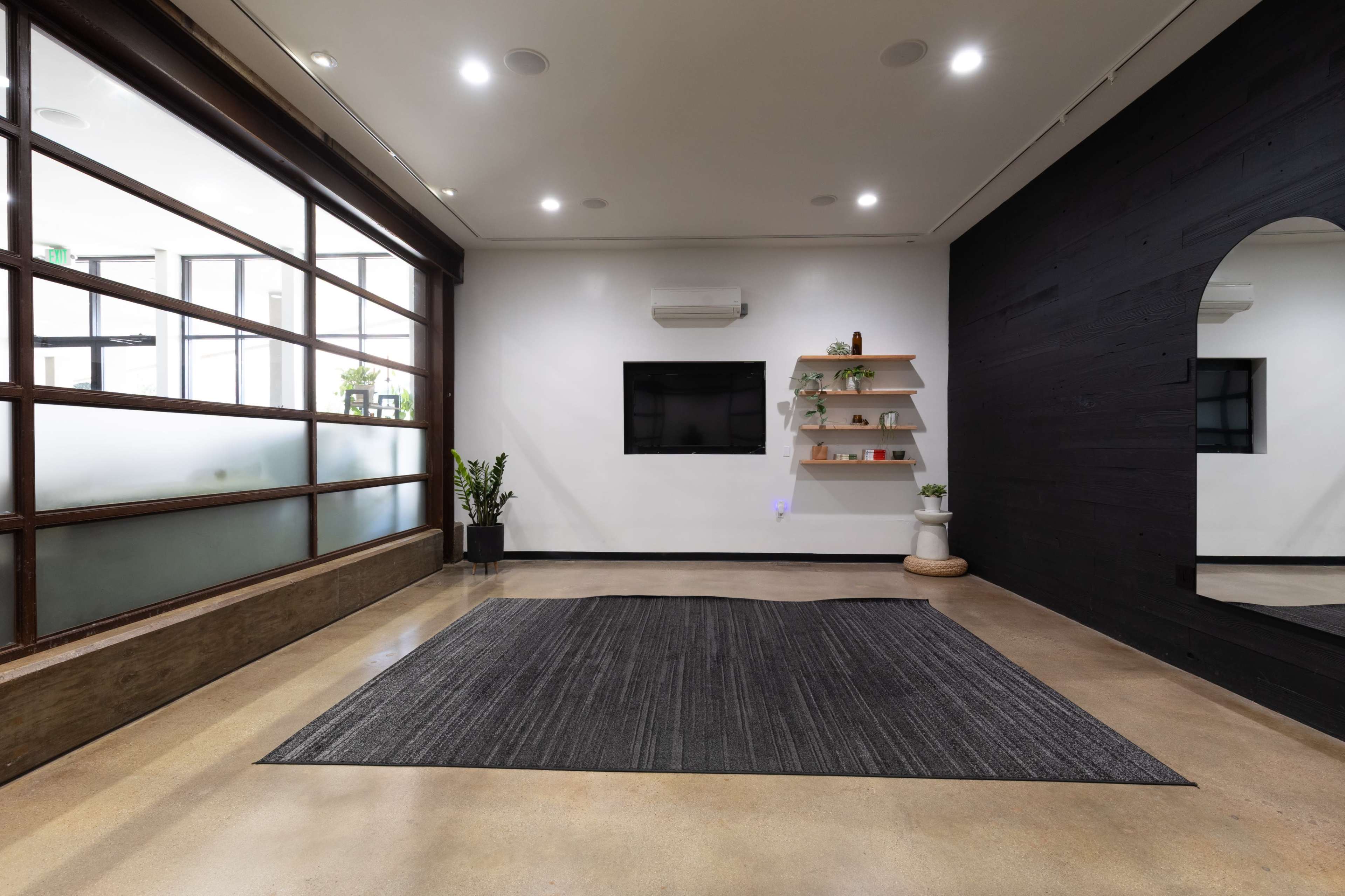 The room features a large area rug on a polished floor, with a black accent wall, a wall-mounted television, and decorative shelves against a light wall.