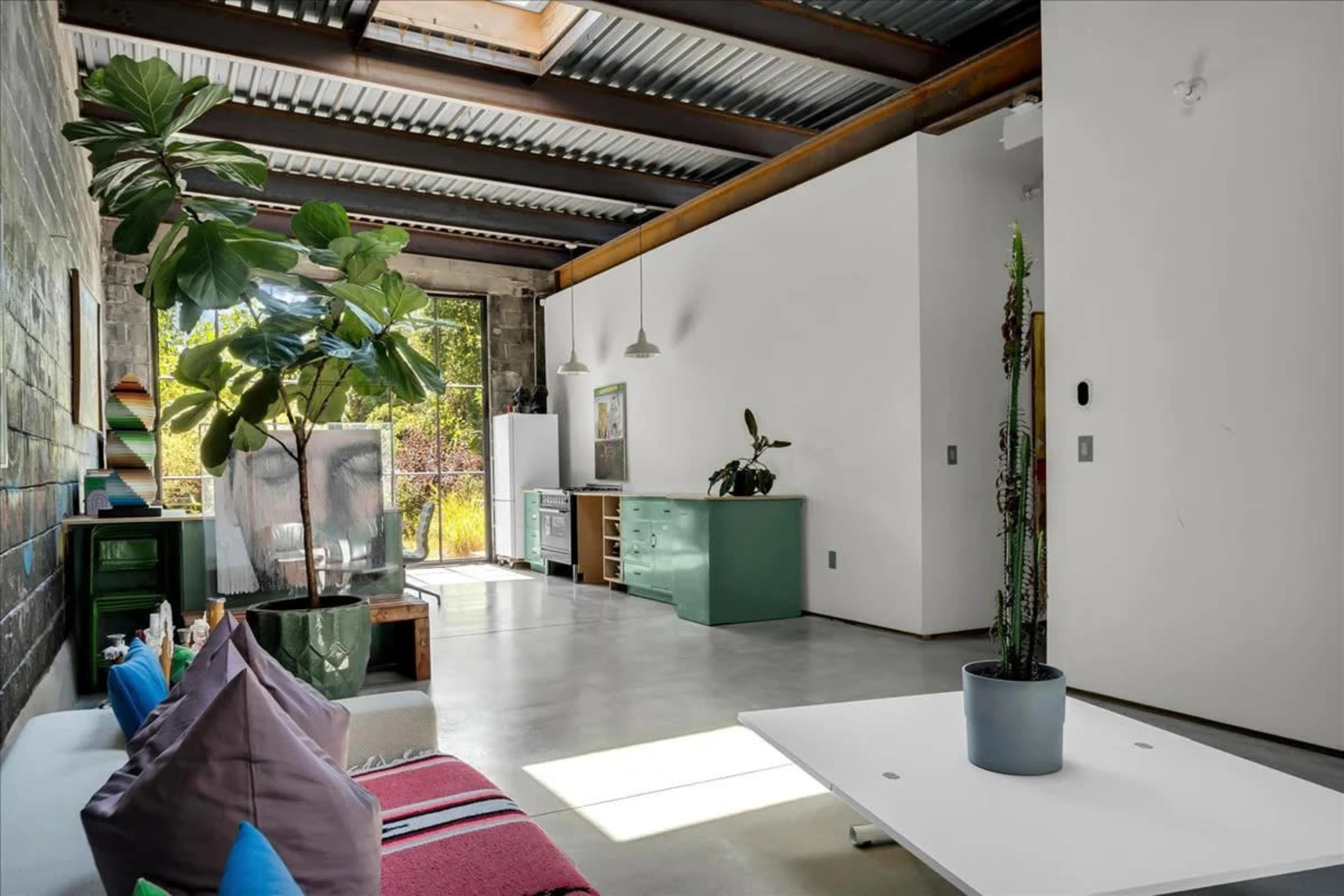The image shows a modern, open-concept living space with concrete floors, large windows, and minimalistic furniture, including a sofa, coffee table, and plants.