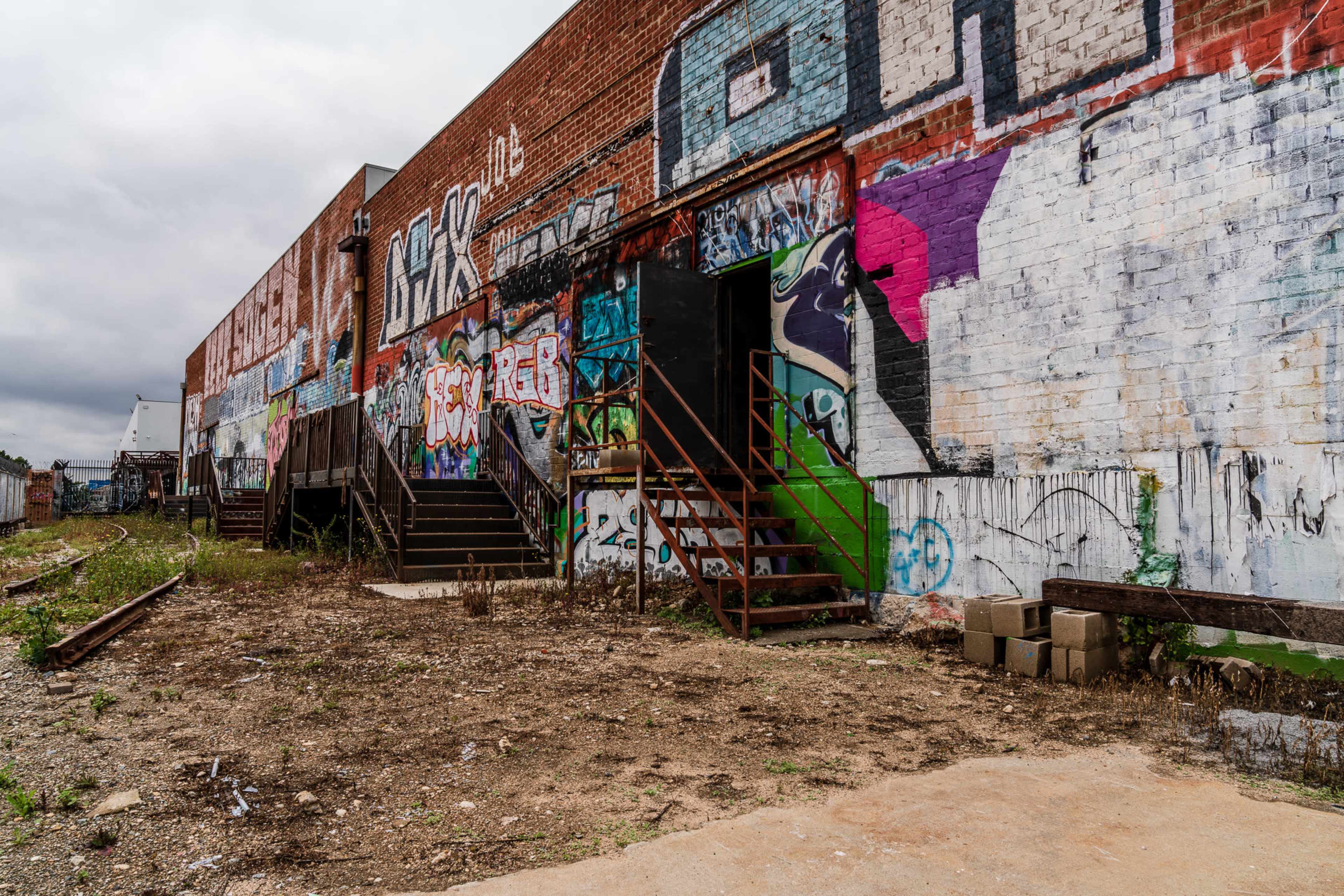A brick building with colorful graffiti on its exterior, featuring a set of metal stairs leading to an entrance and overgrown weeds in the surrounding area.