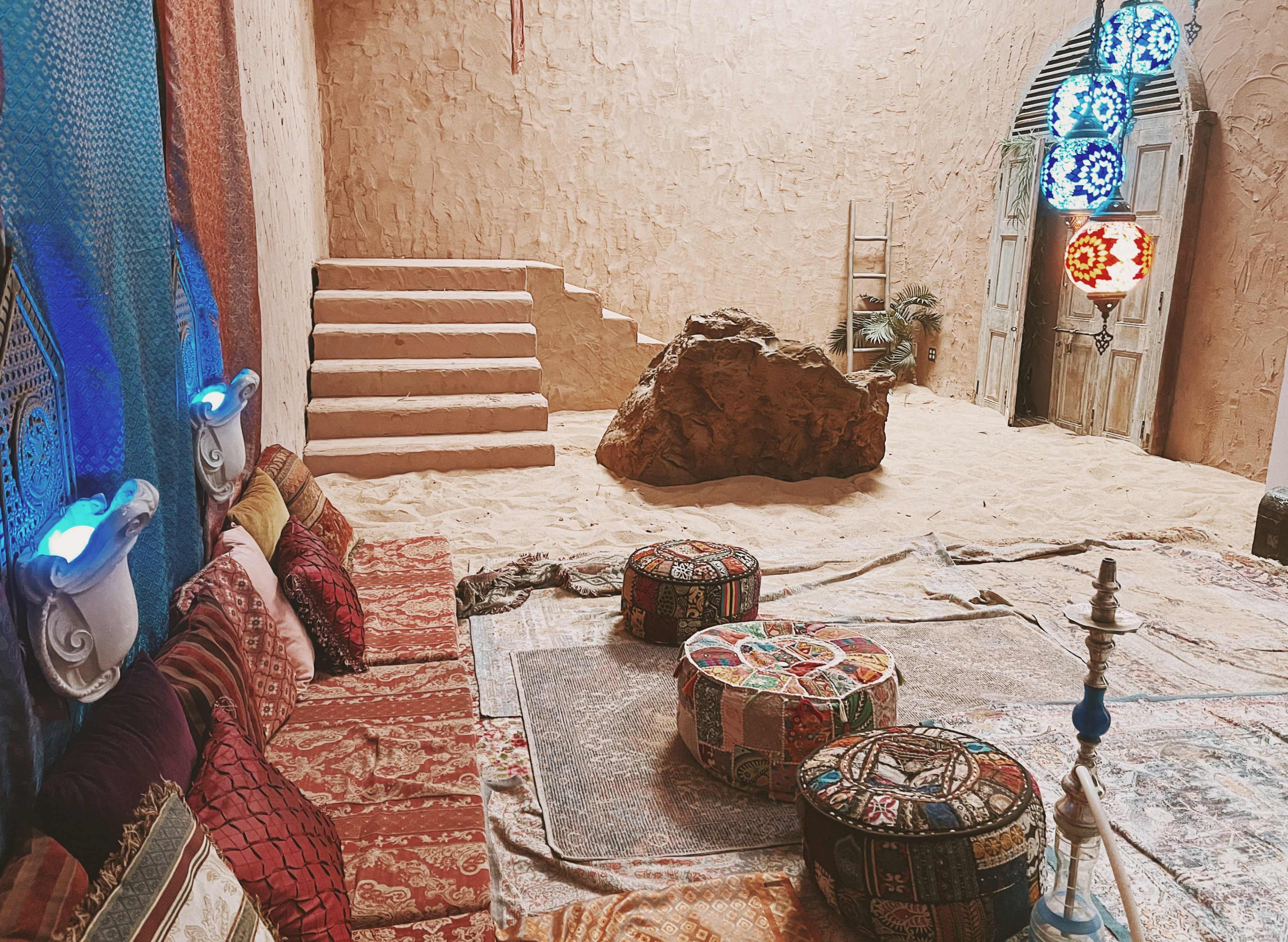 The interior of a desert-themed room features sandy flooring, decorative cushions along a wall, a large rock centerpiece, and colorful lanterns hanging from the ceiling.