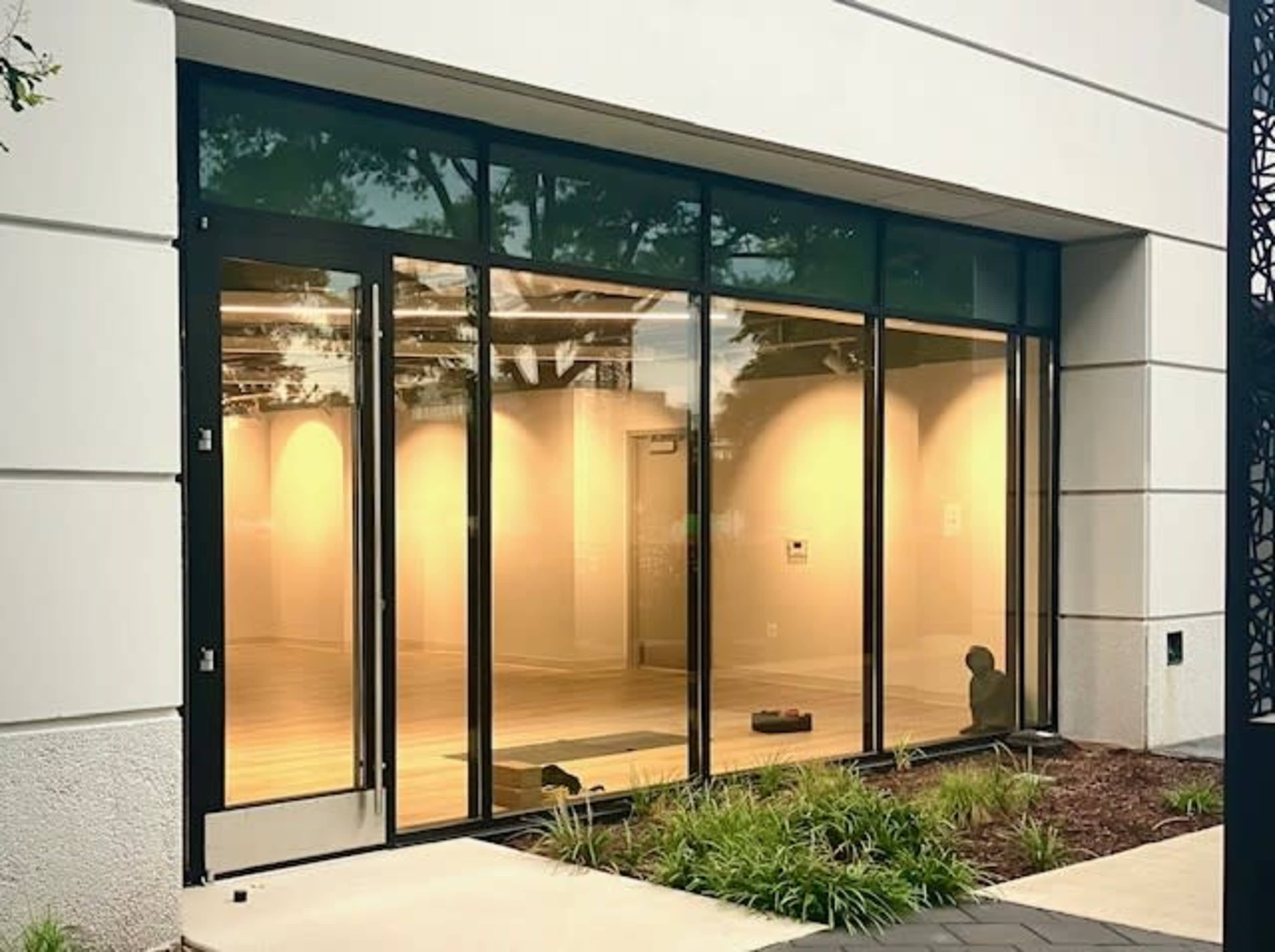 The image shows a modern storefront with large glass windows and doors, revealing an empty interior space with wooden flooring and neutral-colored walls.