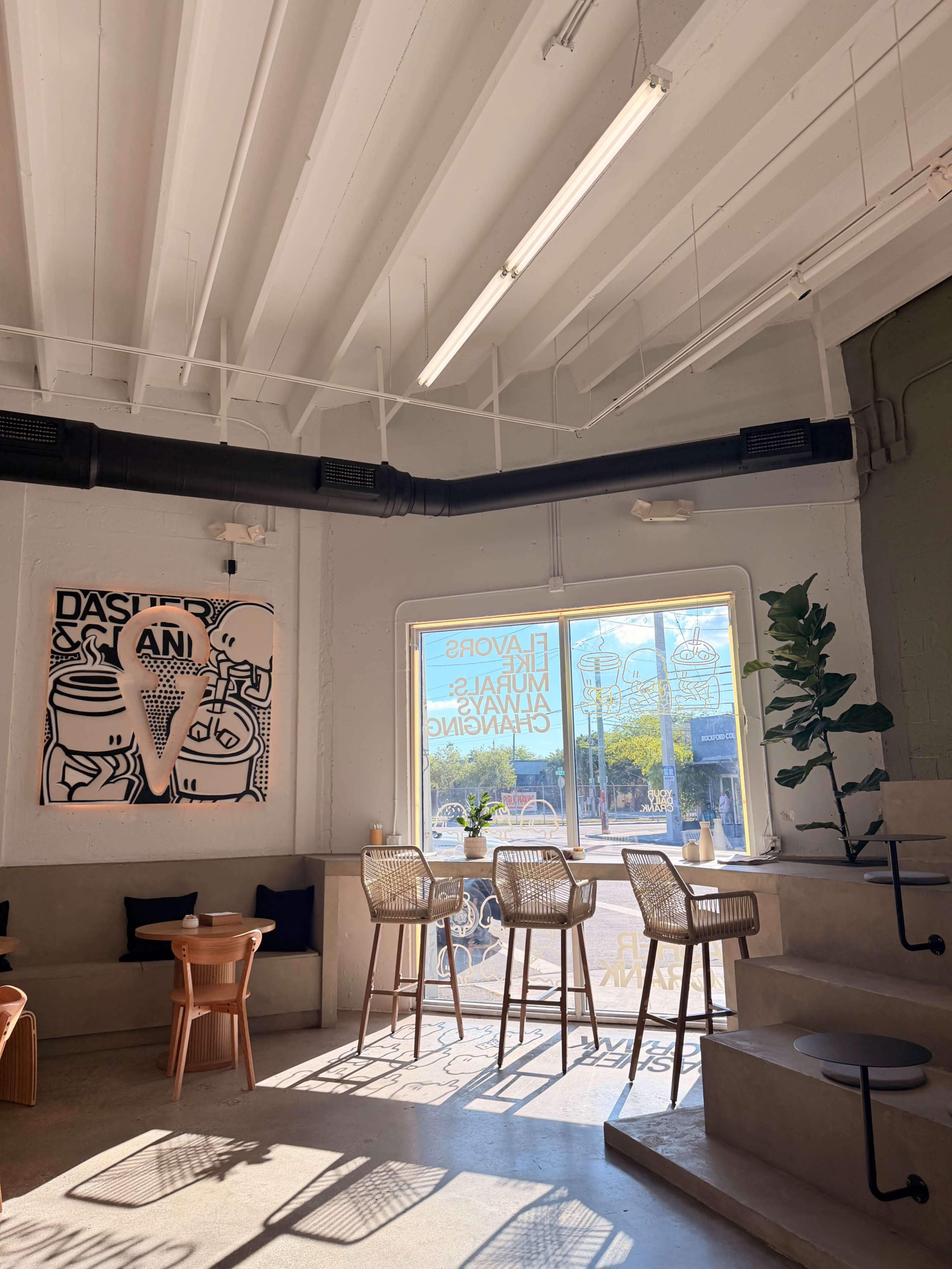 The image shows a bright café interior with seating arrangements, a large window, and a decorated wall featuring an artwork.