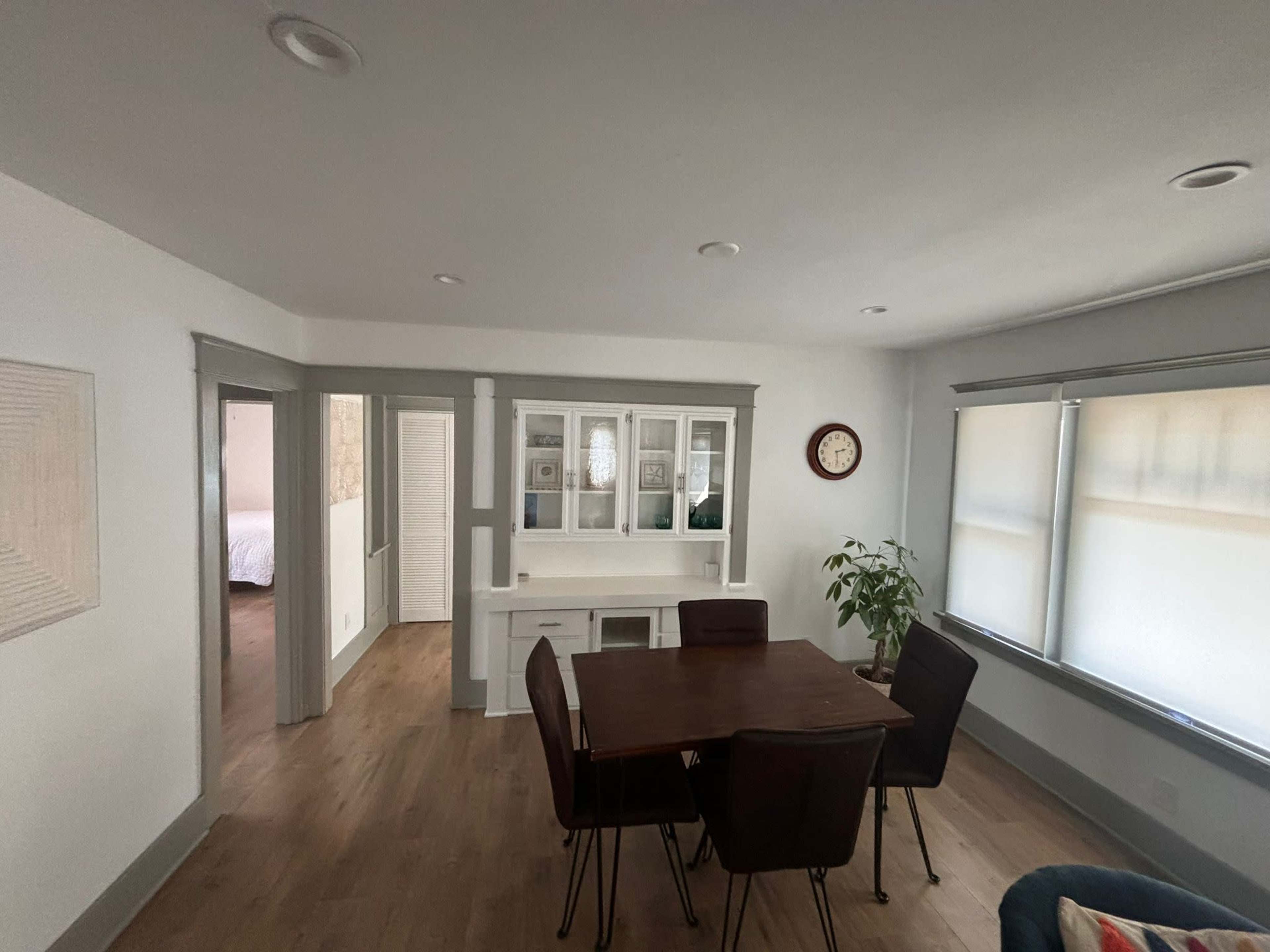 The image shows a bright, spacious room with a dining table and chairs, a cabinet with glass doors, and large windows allowing natural light.