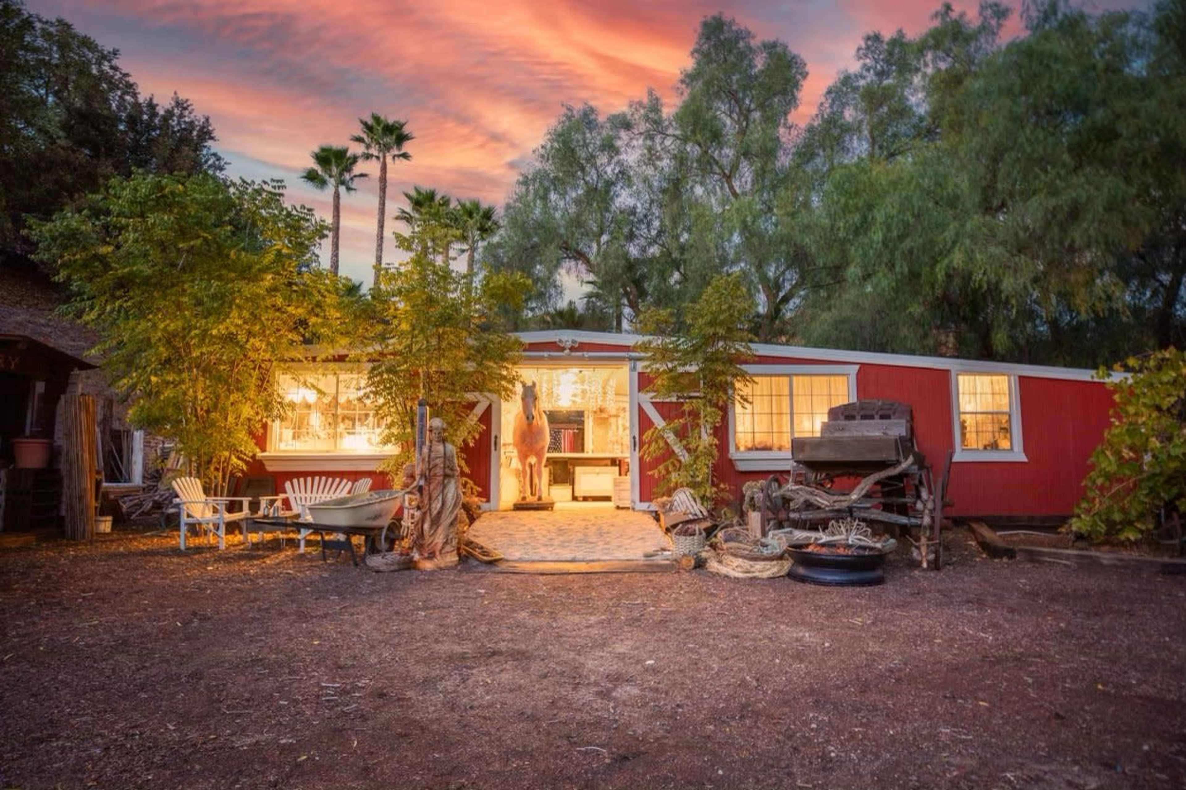 A red cabin with large windows is surrounded by lush greenery and palm trees under a colorful sunset sky.