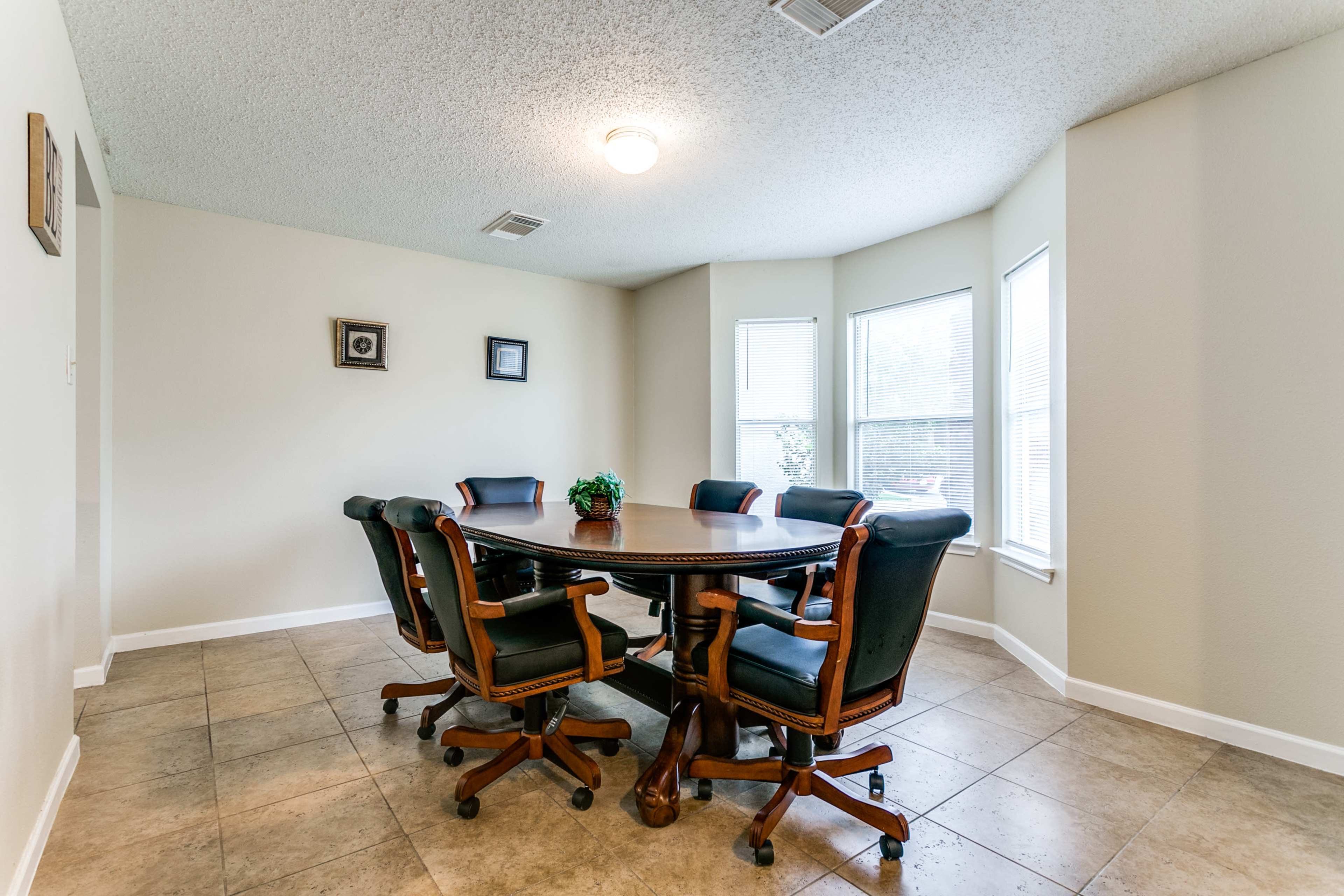 A round wooden table surrounded by six black and brown office chairs is set in a well-lit corner of a room with large windows.
