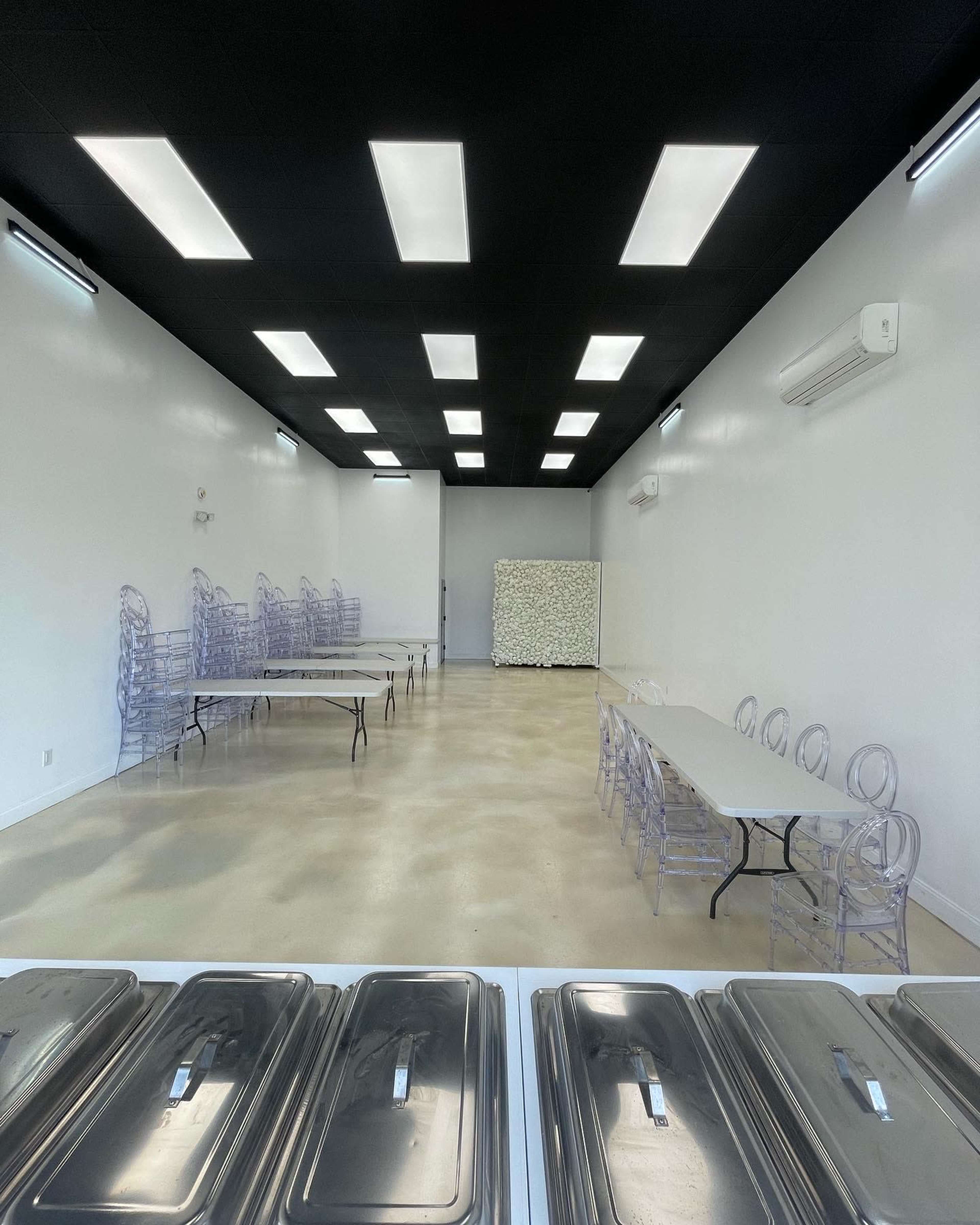 An empty room with white walls, a black ceiling, several clear plastic chairs stacked against the wall, and long tables set up with food trays in the foreground.