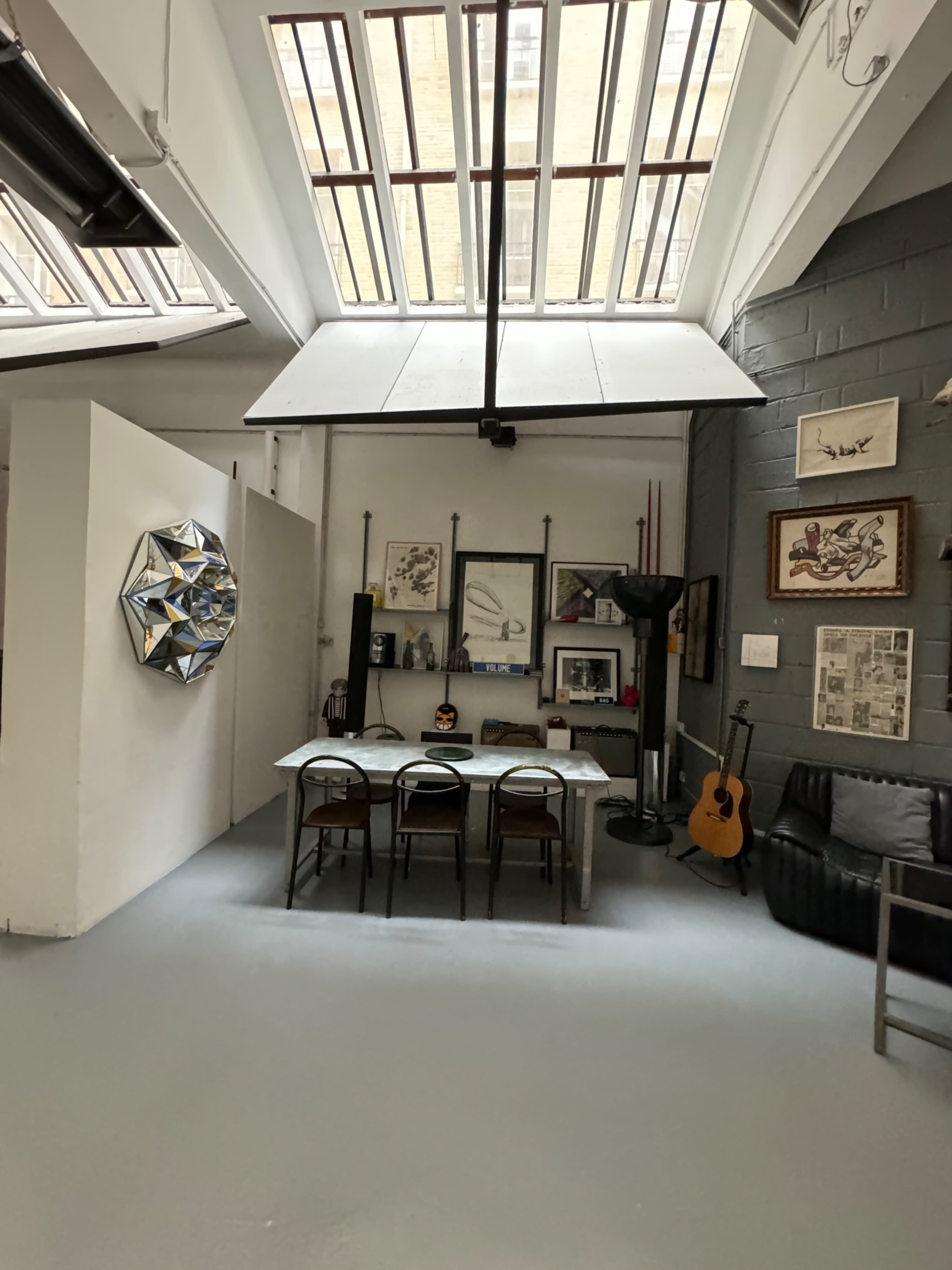 The image shows a spacious studio with a large skylight, featuring a table with chairs, artistic wall decor, and a guitar in the corner.