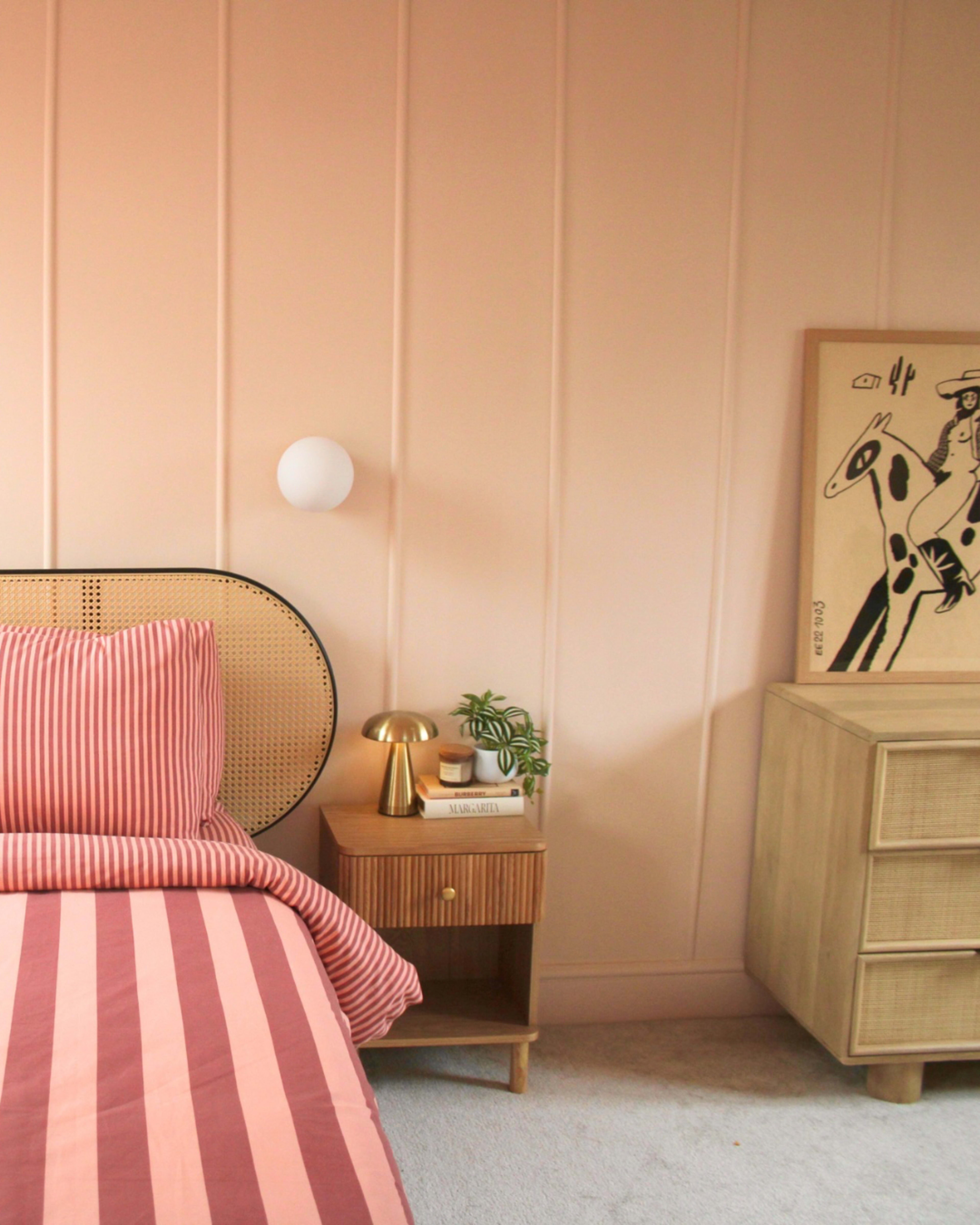The image shows a bedroom with a striped bedspread, a bedside table with a lamp and a small plant, and a piece of artwork on the wall.