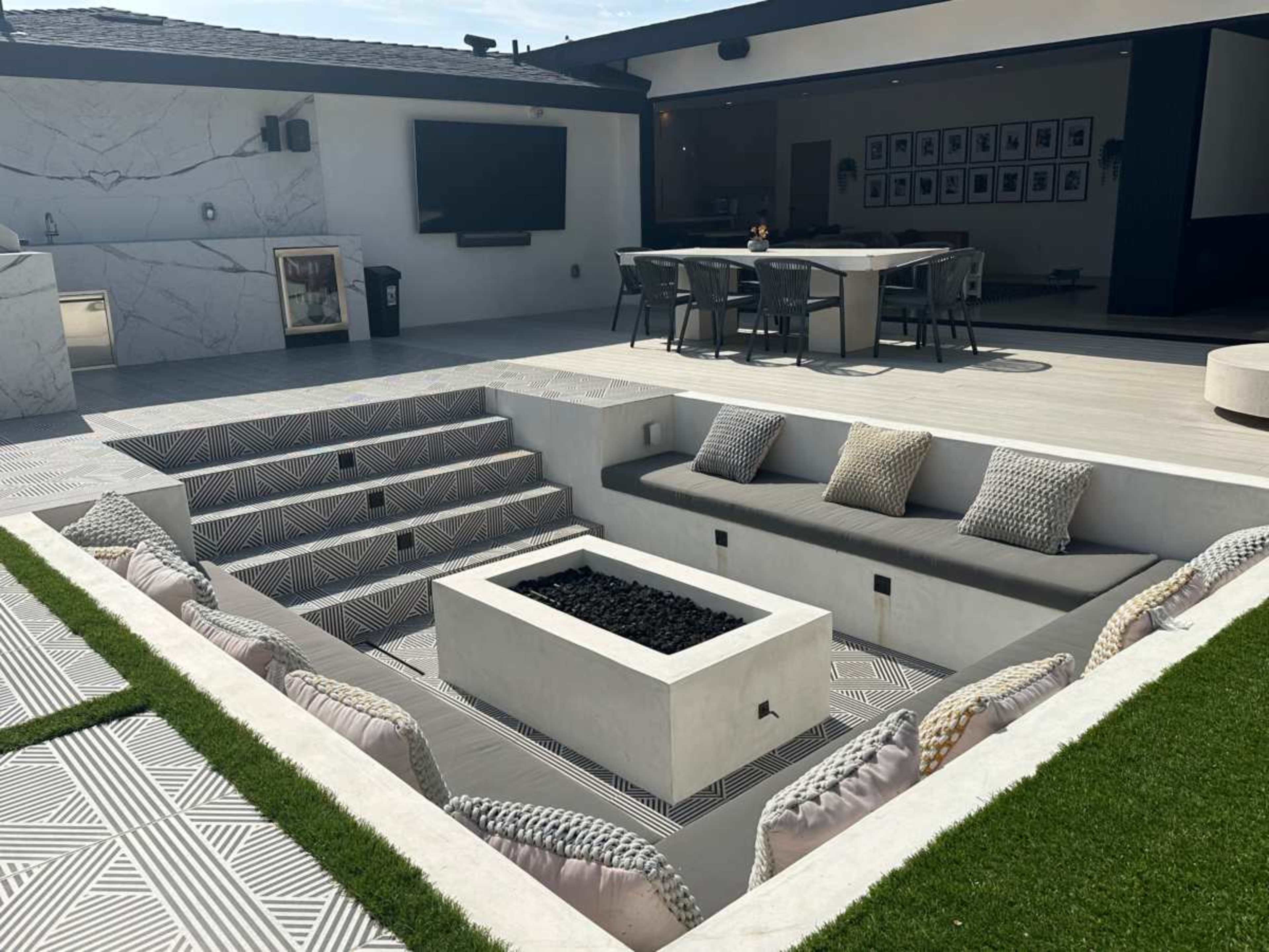 The image shows a modern outdoor living area featuring a sunken seating area with a fire pit, surrounded by steps and a patio leading to an open kitchen and dining space.