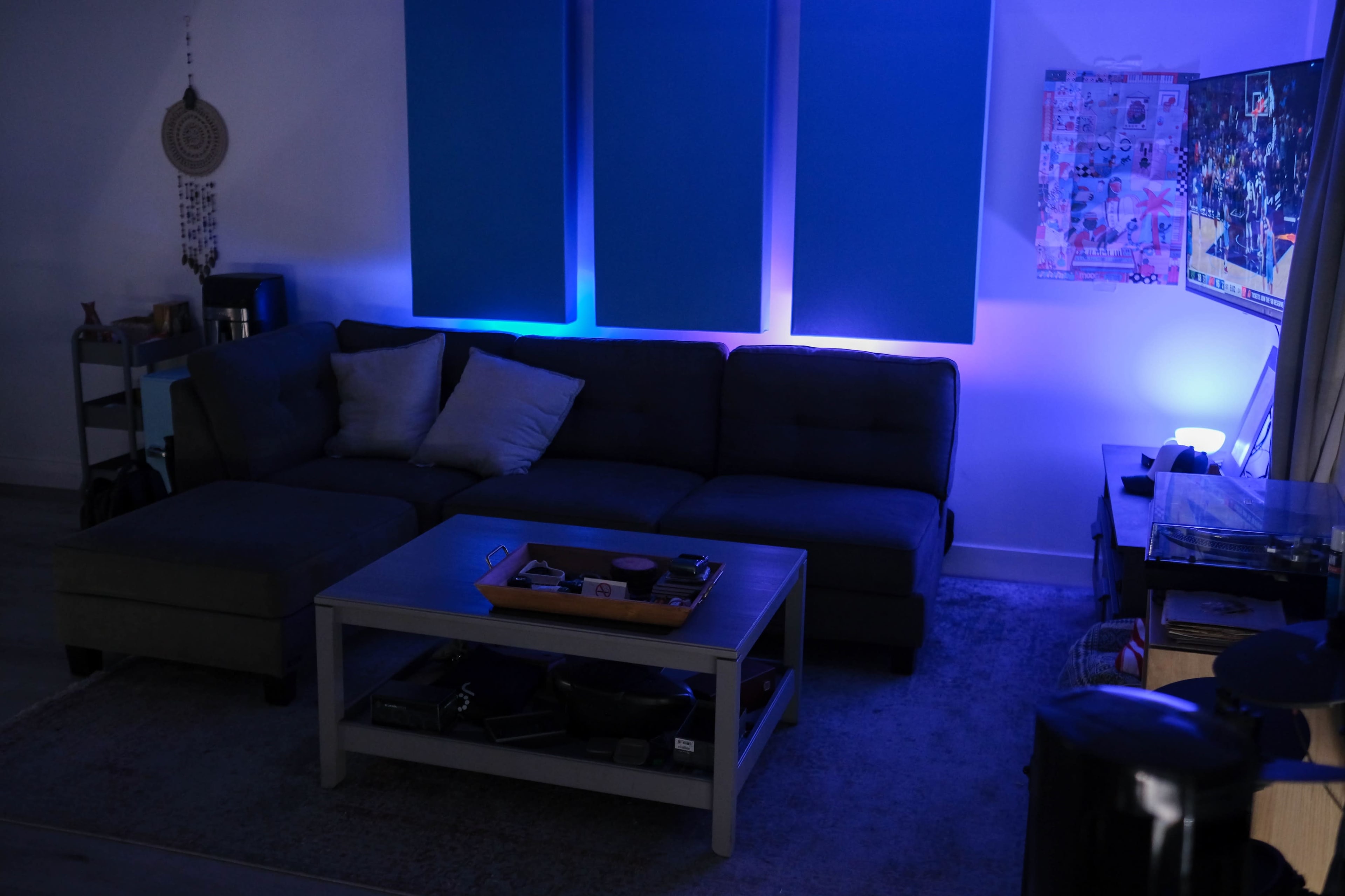 A modern living room features a sectional couch with cushions, a coffee table, and blue wall panels, illuminated by soft lighting.