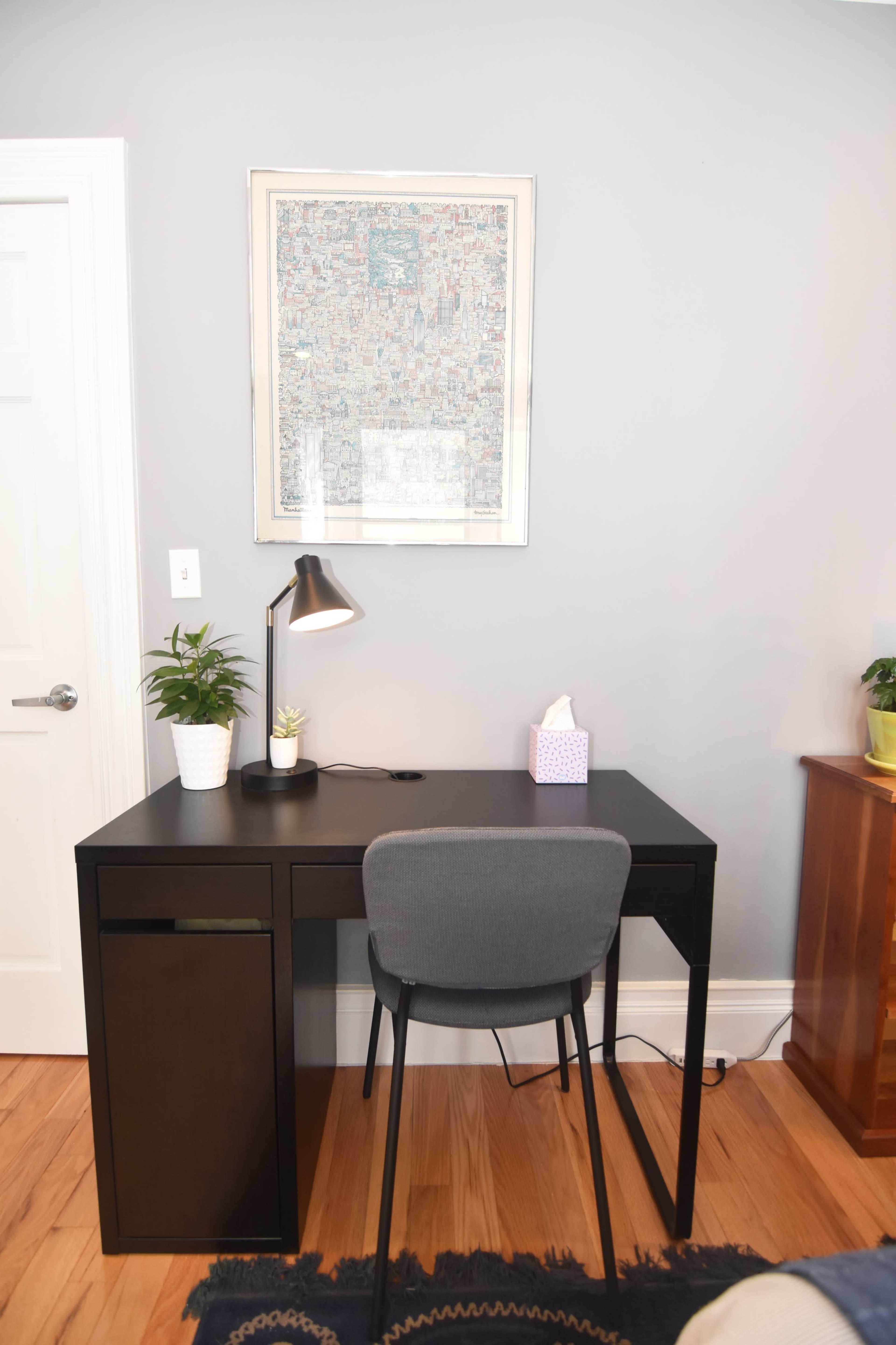 A black desk with a lamp, a small plant, and a tissue box is positioned against a gray wall, beneath a framed piece of artwork.