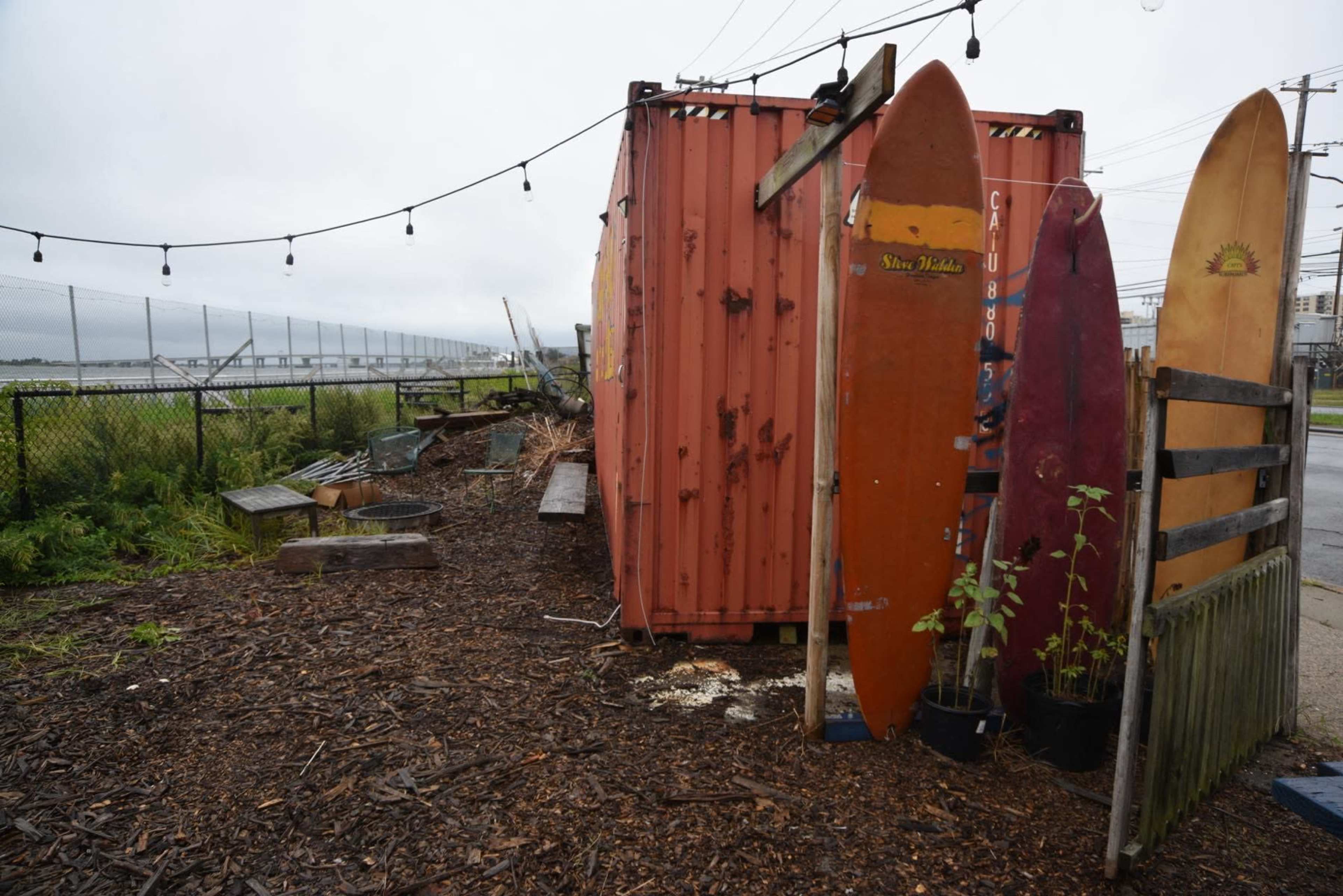 A weathered shipping container next to several surfboards and a landscaped area with wood chips and potted plants.