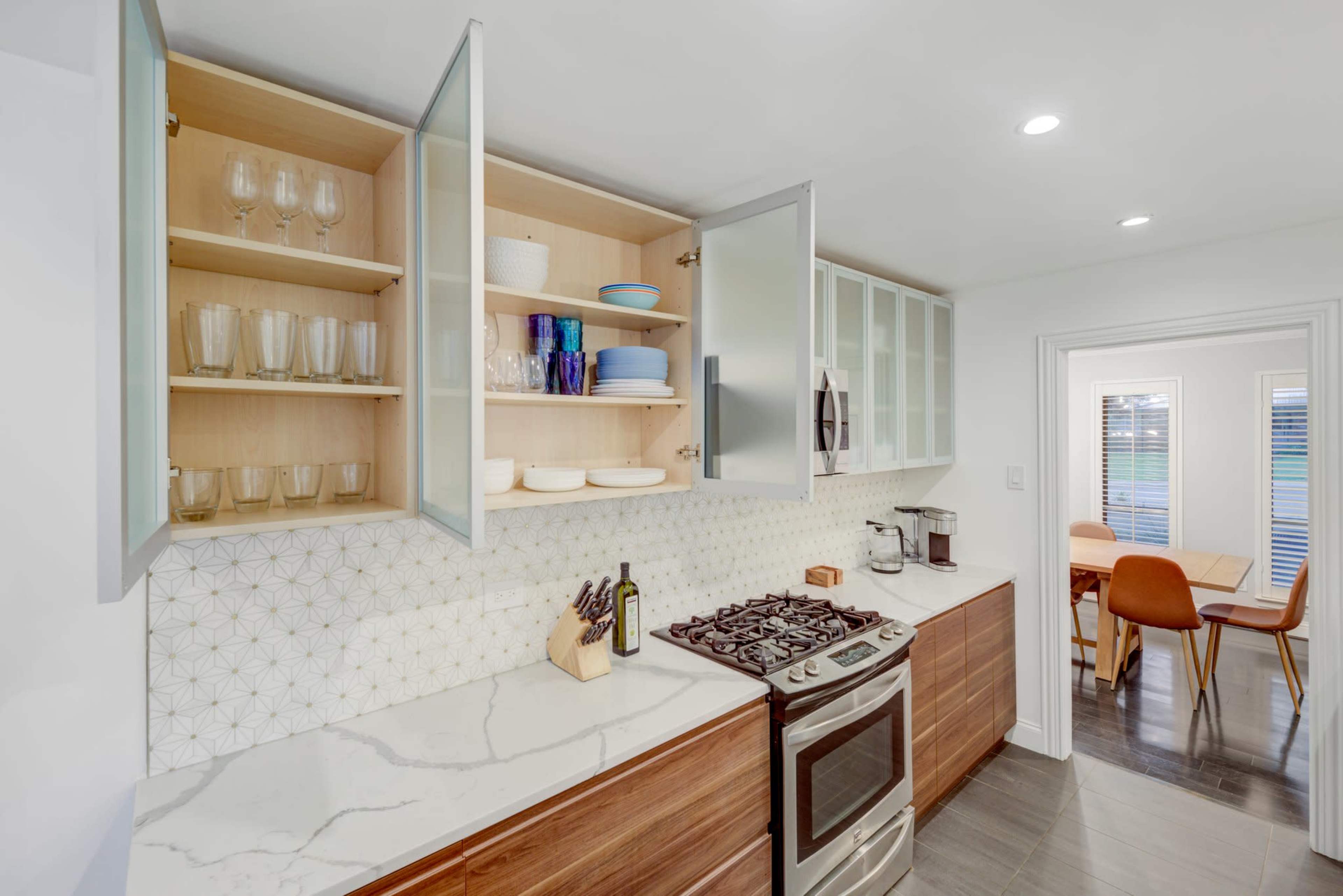 A modern kitchen features open shelves displaying glassware and dishes, with a gas stove and a dining area visible in the background.