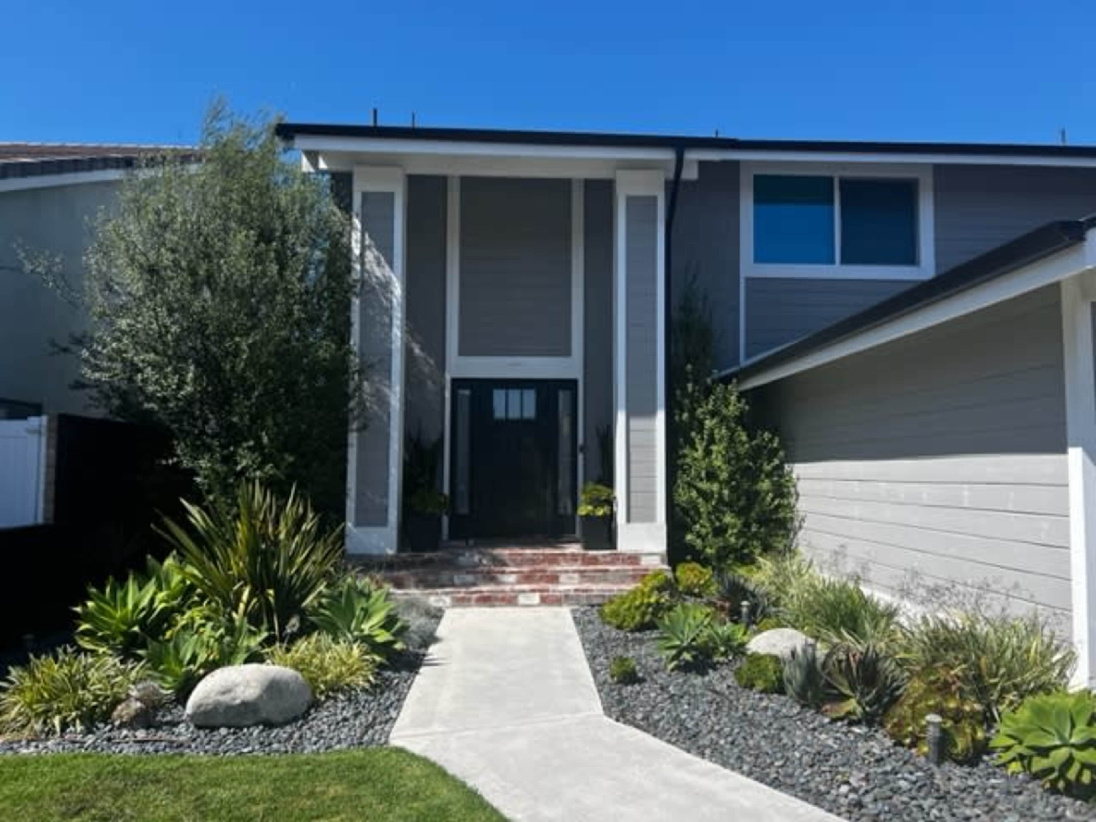 The image shows a modern house with a prominent entrance flanked by landscaped greenery and stone pathways.