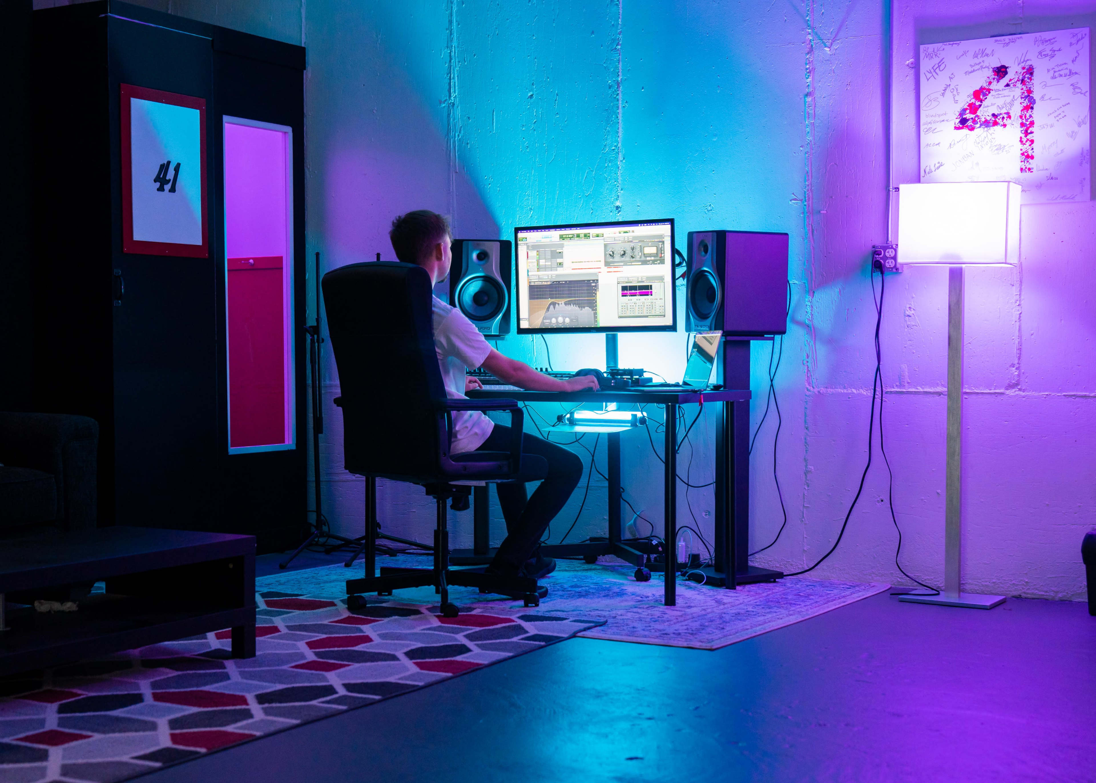 A person sits at a desk in a room with blue and purple lighting, working on a computer surrounded by speakers.