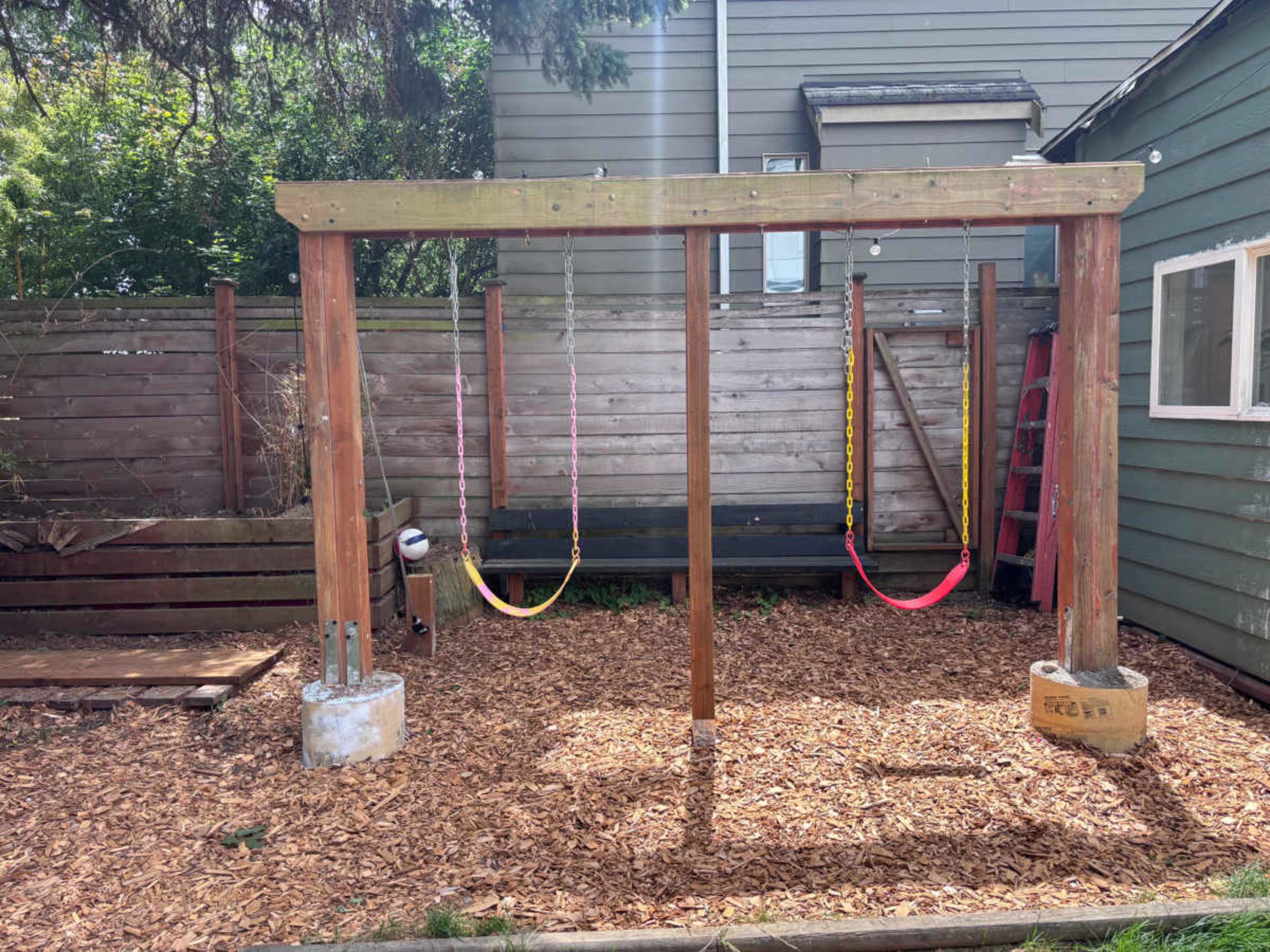 The image shows a wooden swing set with two swings, set on a bed of wood chips in a fenced backyard.