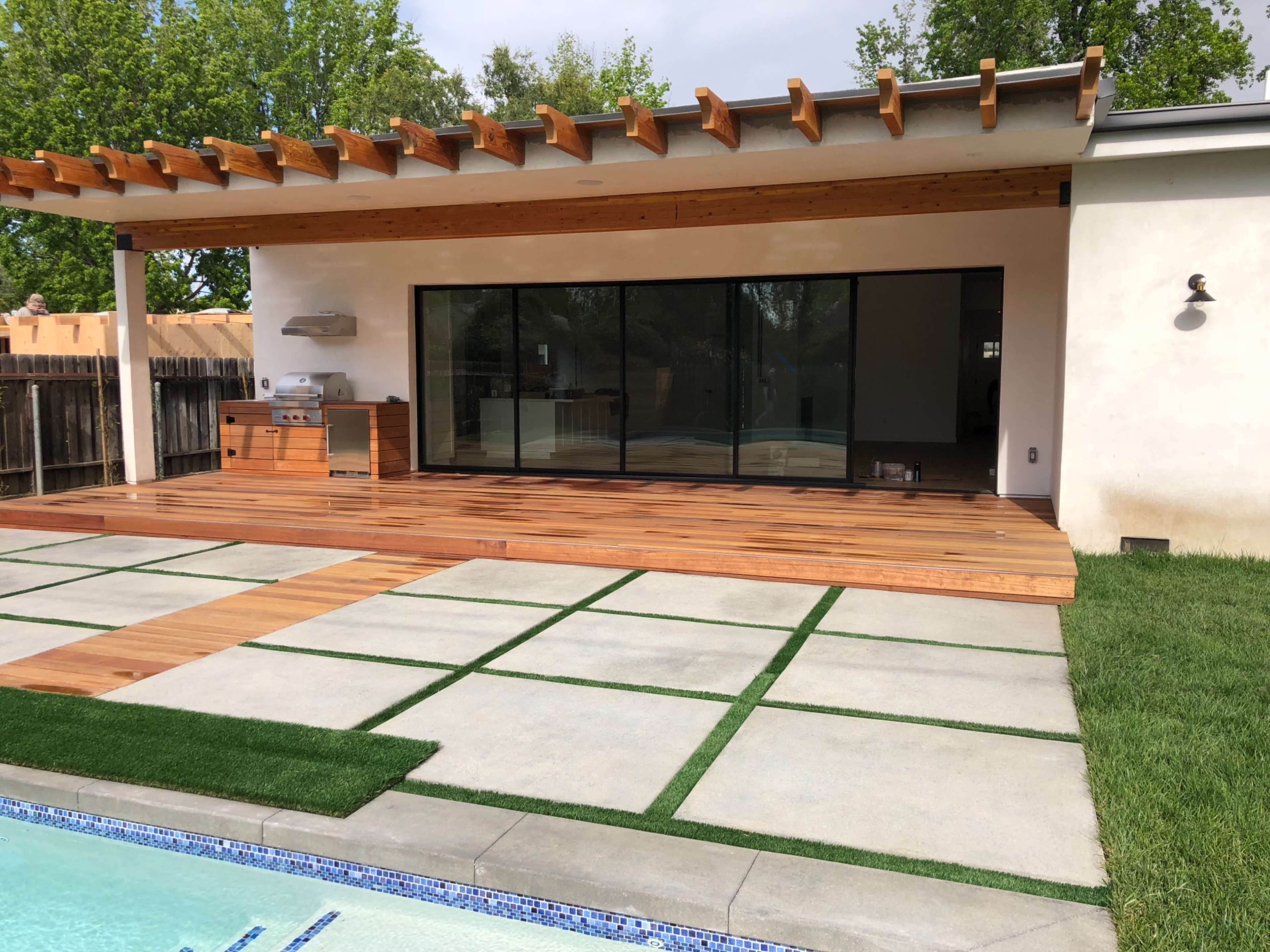A modern backyard with a wooden deck, a glass sliding door, and concrete paving stones bordered by grass.