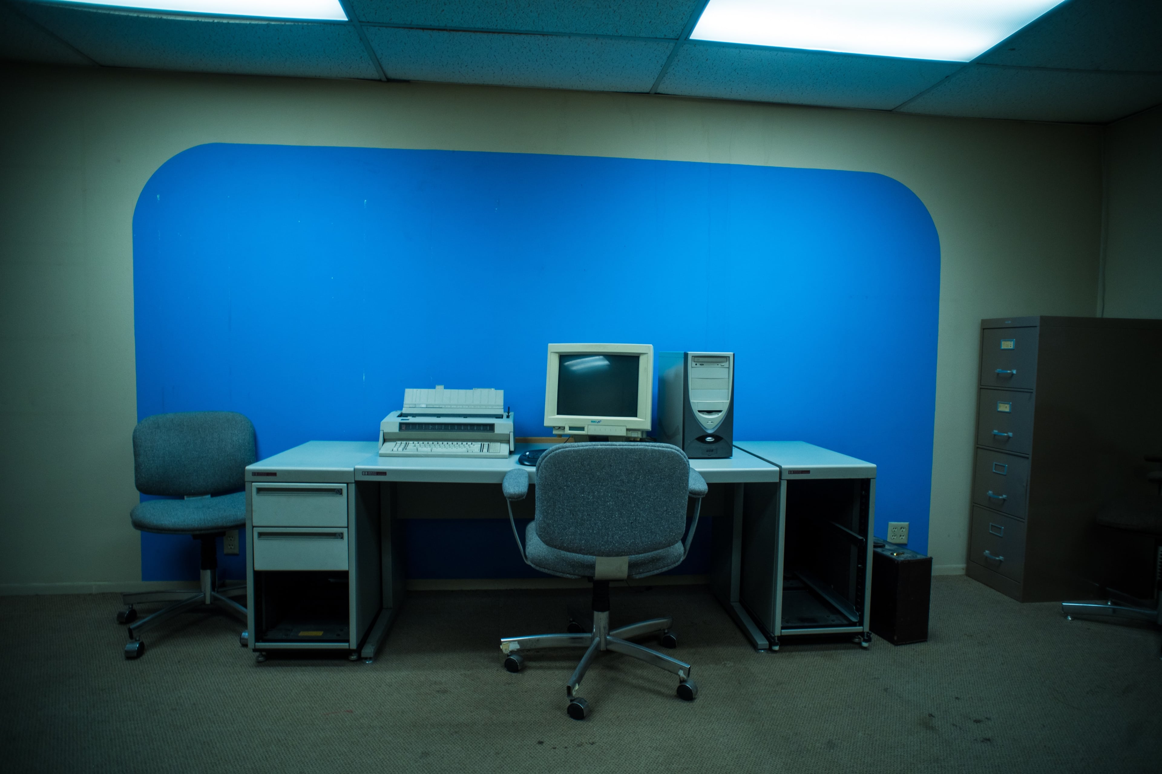 An empty office space features a desk with a computer monitor, a printer, and two chairs against a blue wall.