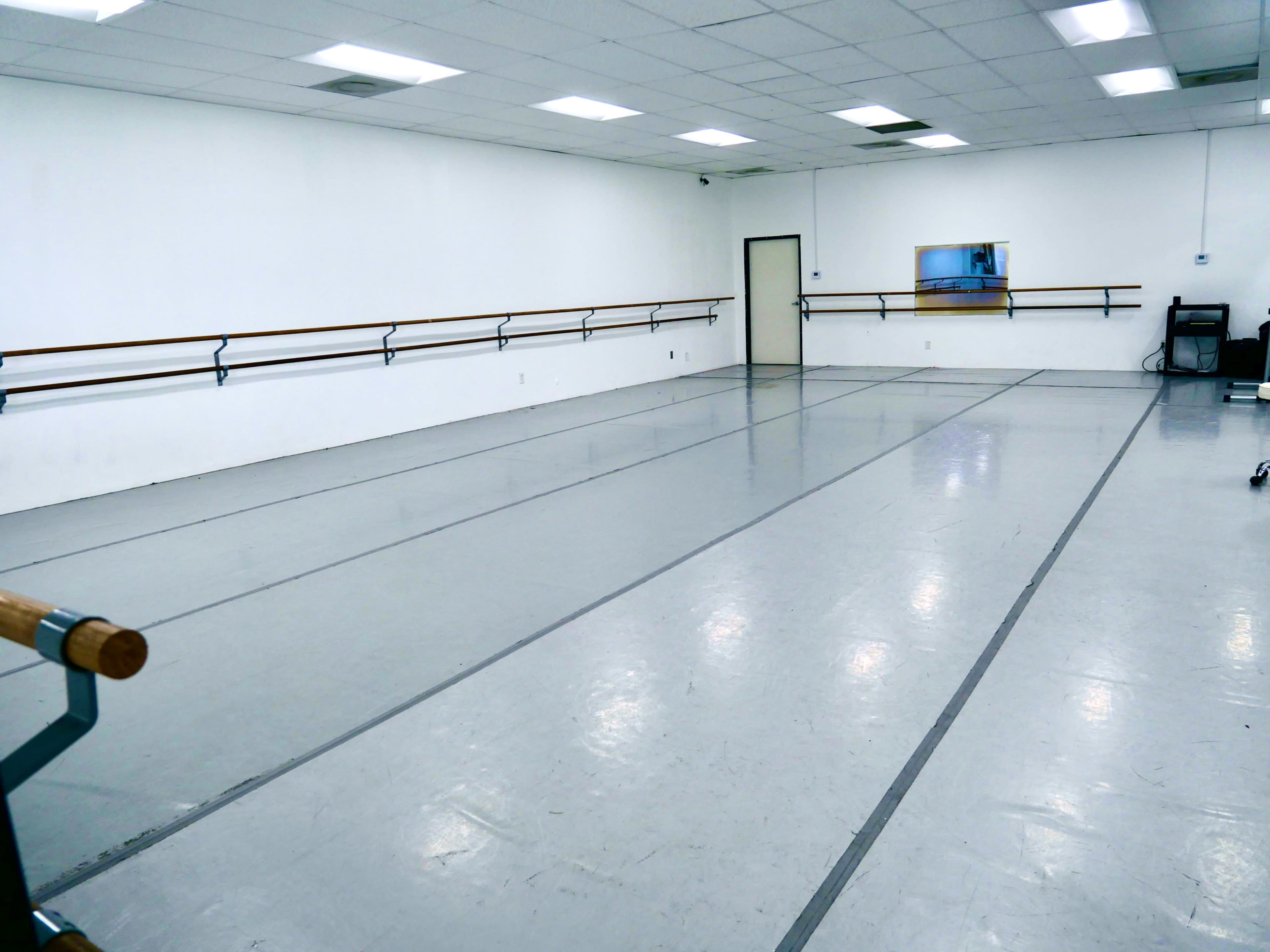 The image shows a spacious dance studio with wooden ballet bars mounted on white walls and a mirrored panel.