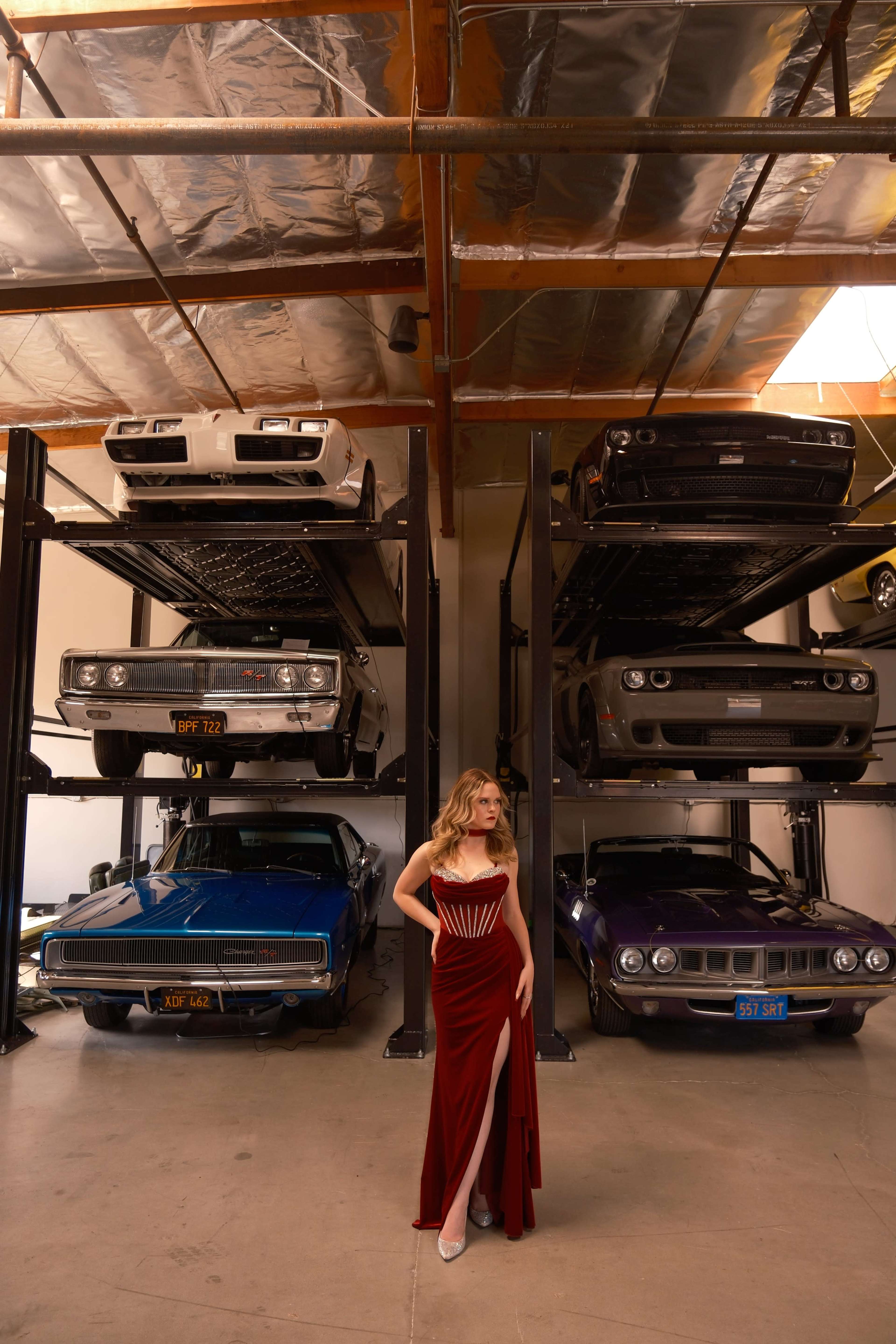 A woman in a red dress stands in a garage filled with classic cars displayed on multiple levels.