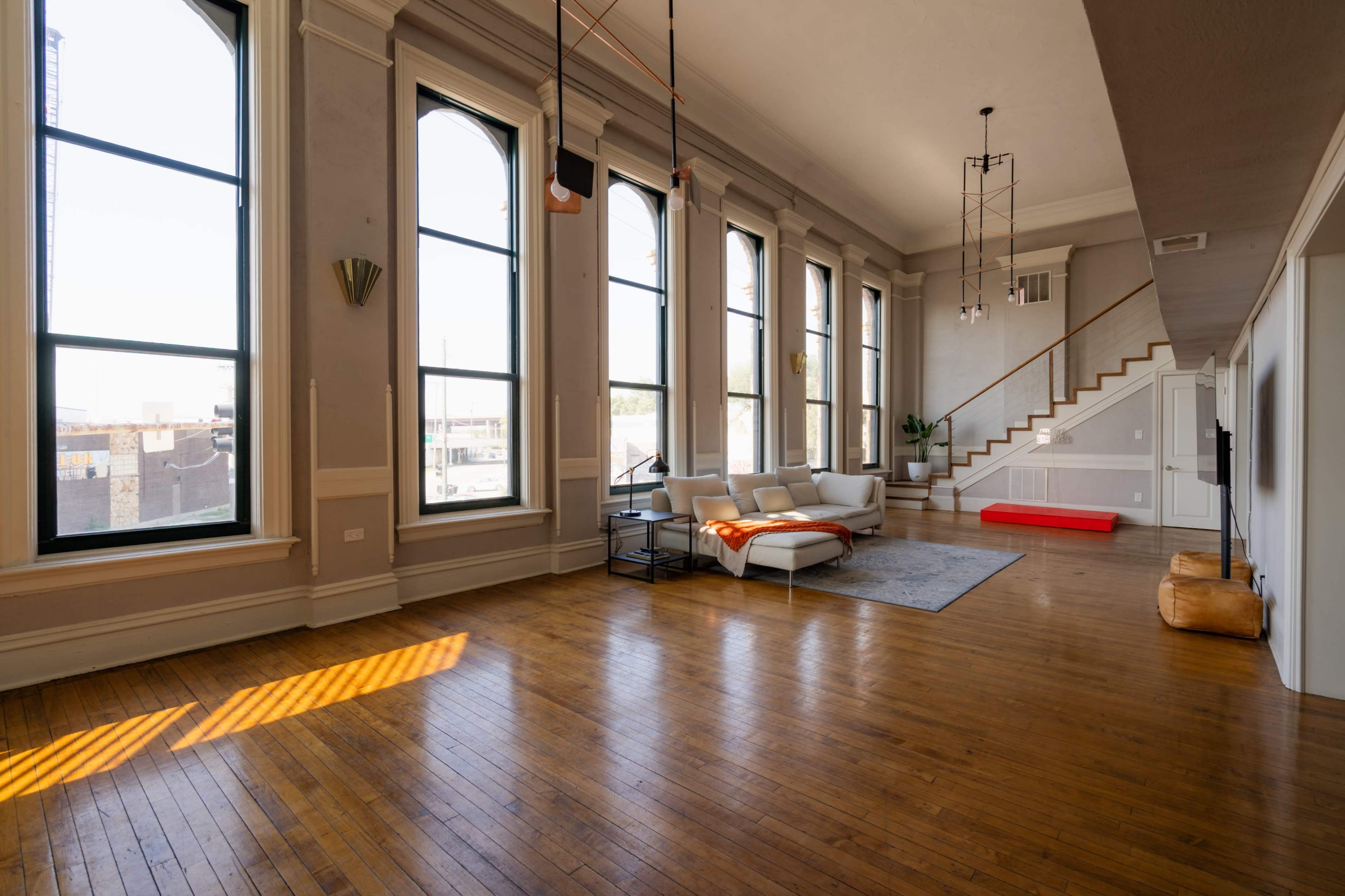 The image shows a spacious living area with large windows, hardwood floors, and a modern sofa arrangement.