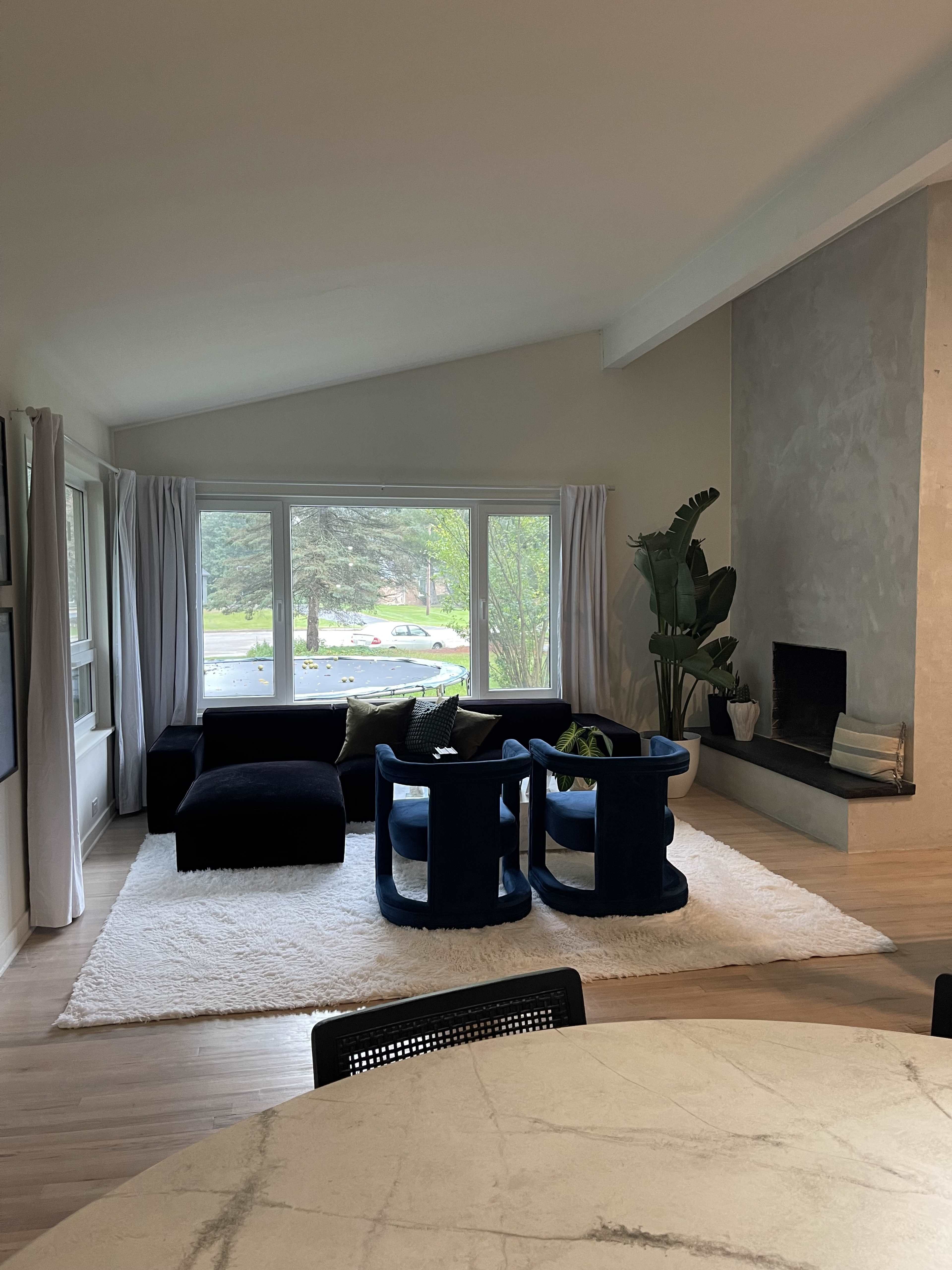A modern living room features a dark blue sectional sofa and two circular chairs arranged on a light rug, with large windows overlooking greenery.