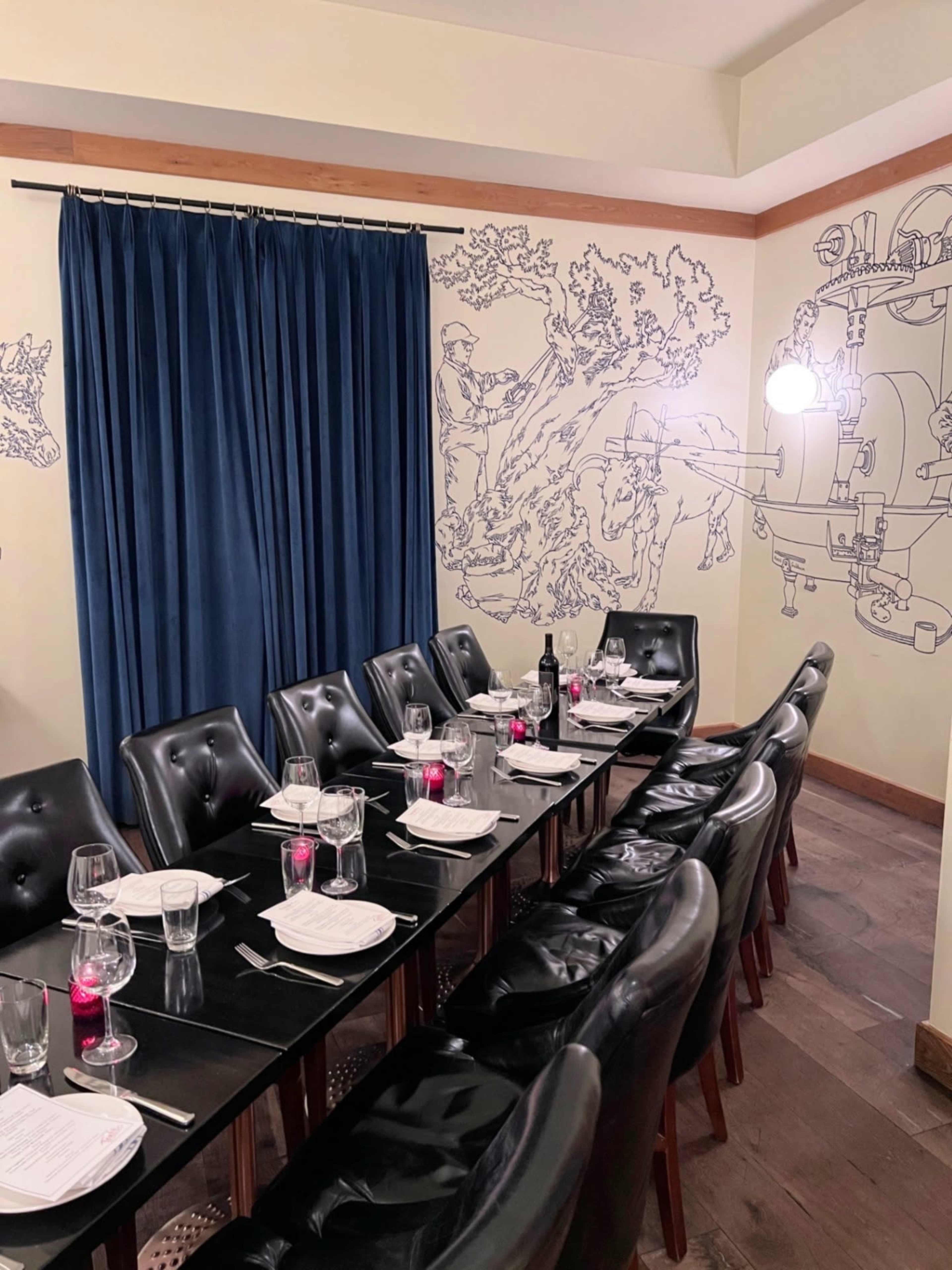 A long table set for dinner is placed in a restaurant, surrounded by artistic wall murals and accented by a dark curtain.