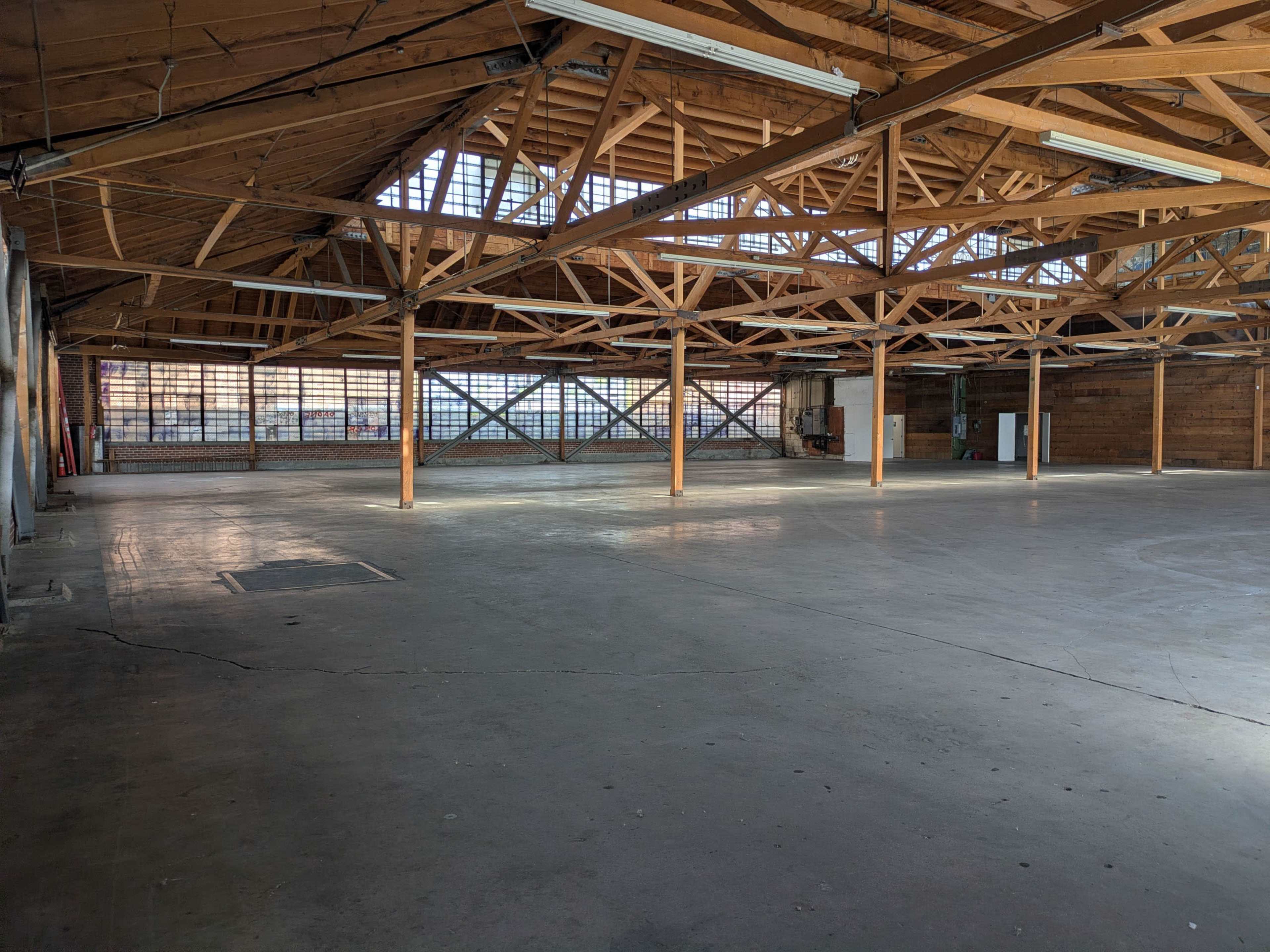 The image shows a large, empty warehouse with exposed wooden beams and concrete flooring, illuminated by natural light from the windows.