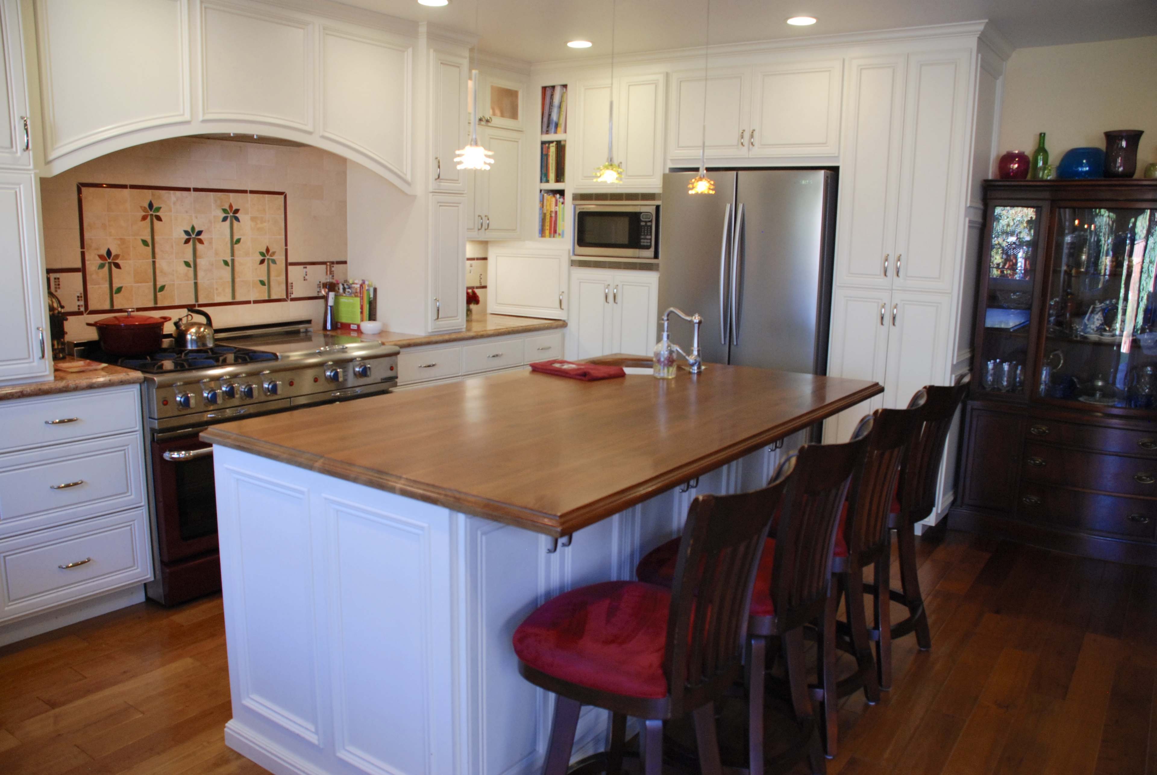 The image shows a modern kitchen with white cabinetry, a central wooden island with four red stools, and stainless steel appliances.