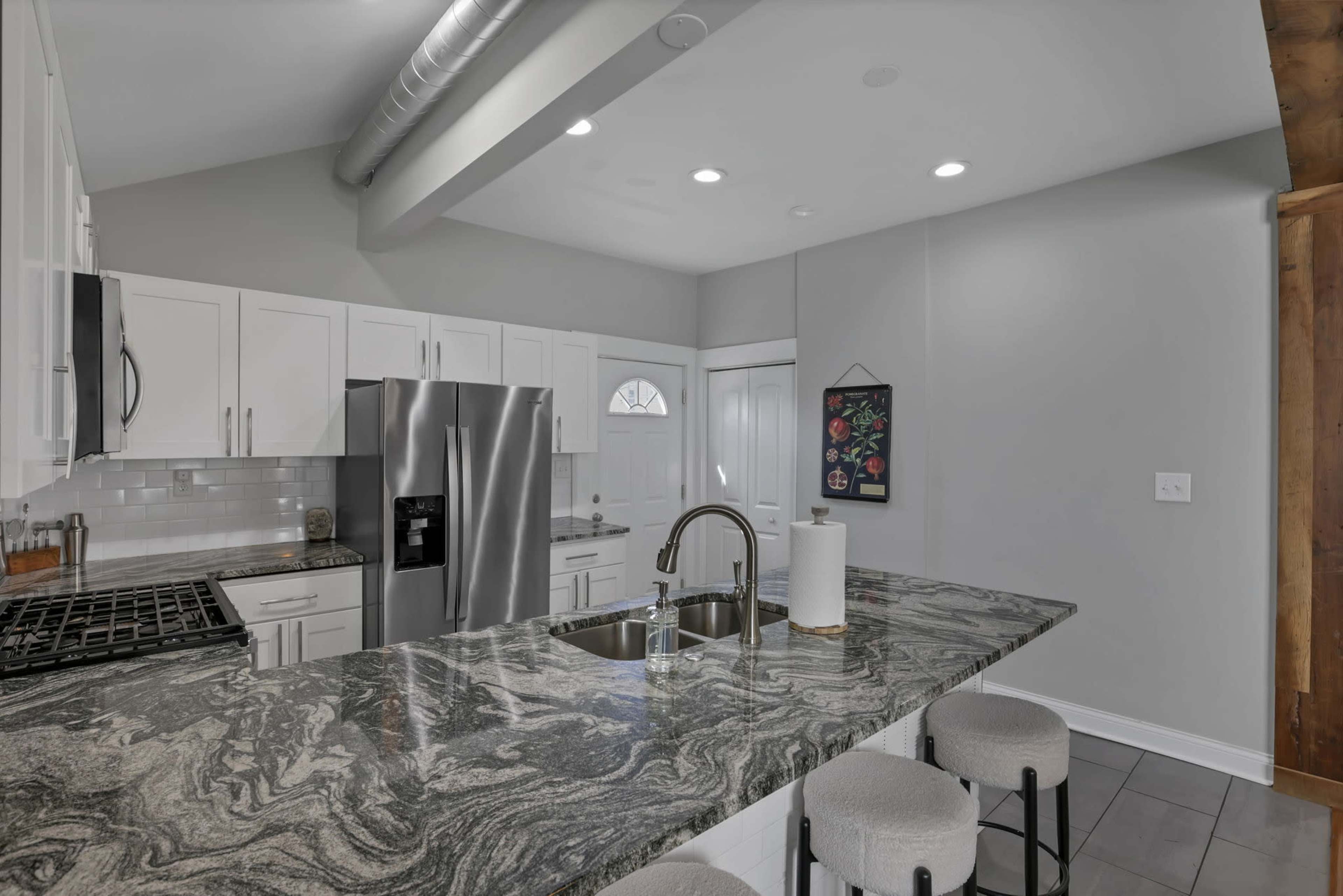 The image shows a modern kitchen with white cabinetry, stainless steel appliances, and a dark granite countertop with seating on one side.