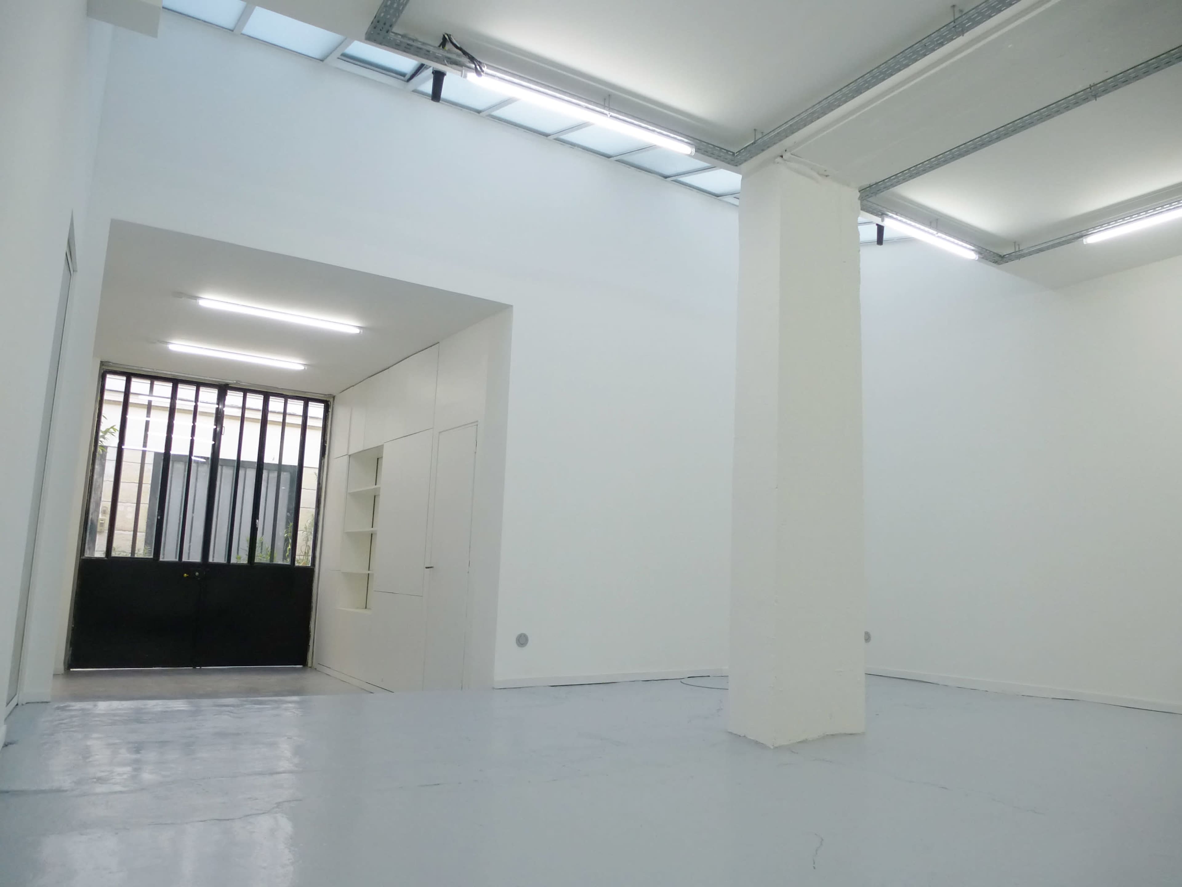 A bright, minimalist interior of a room with white walls and a concrete floor, featuring a doorway with black doors in the background.