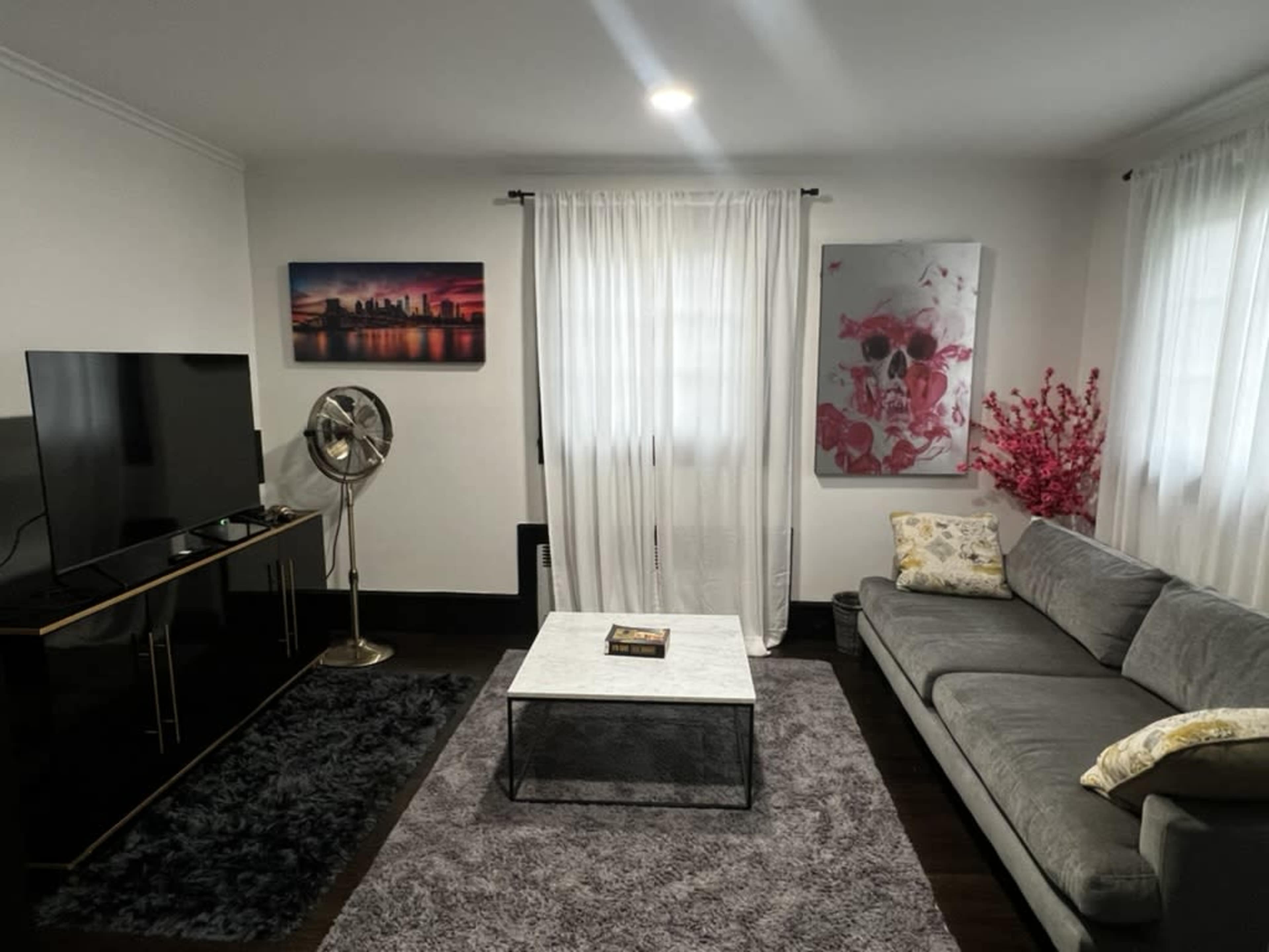 The living room features a gray sectional sofa, a black TV stand with a television, a marble coffee table, and decorative elements including a fan, paintings, and potted plants.
