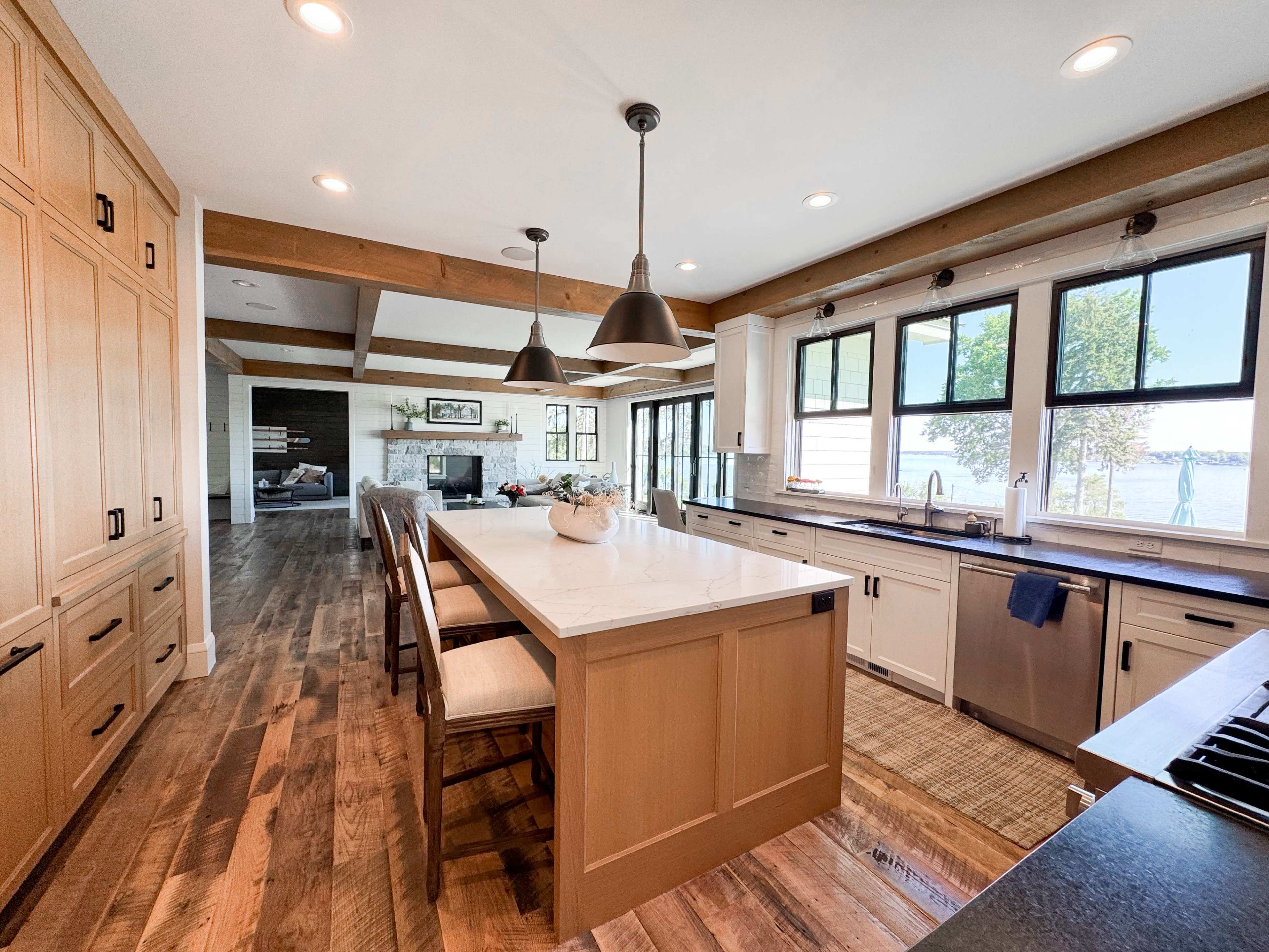A modern kitchen features a large central island, wooden cabinetry, and large windows that overlook a lake.