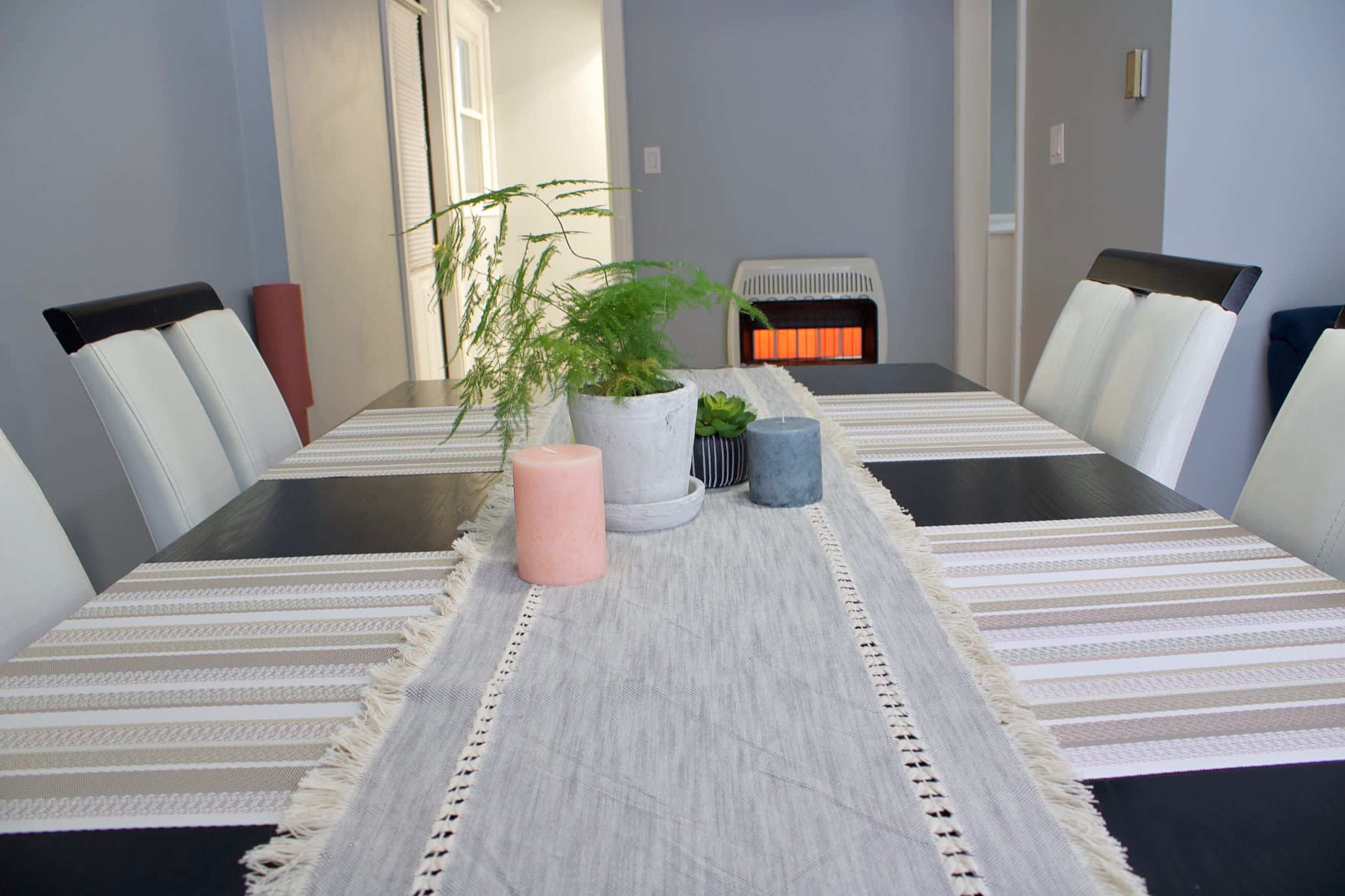 A dining table is set with a table runner, candles, a potted plant, and a small decorative bowl, with a heater visible in the background.