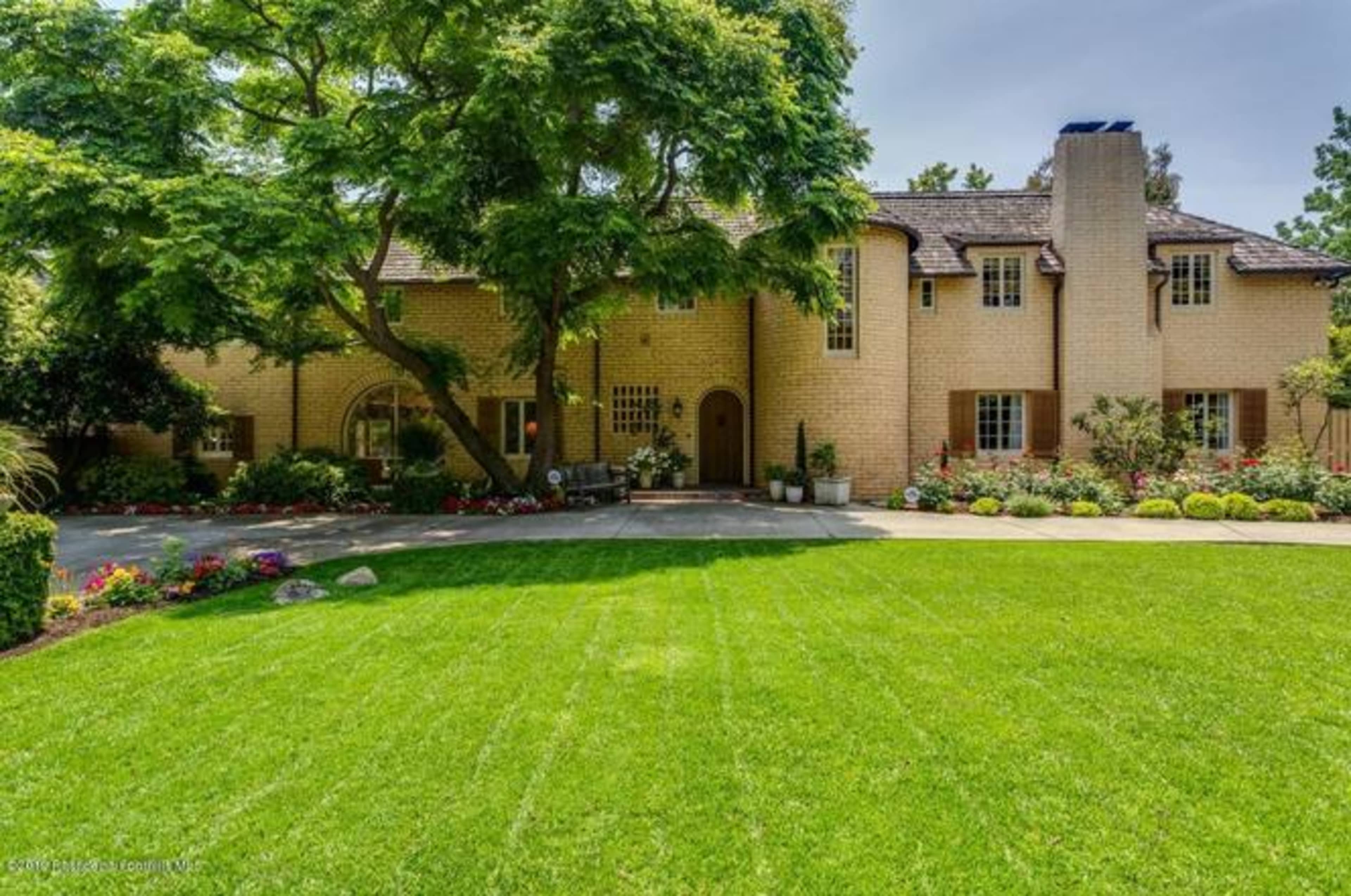 A large brick house with a chimney is set amidst well-maintained lawns and garden beds filled with flowers.
