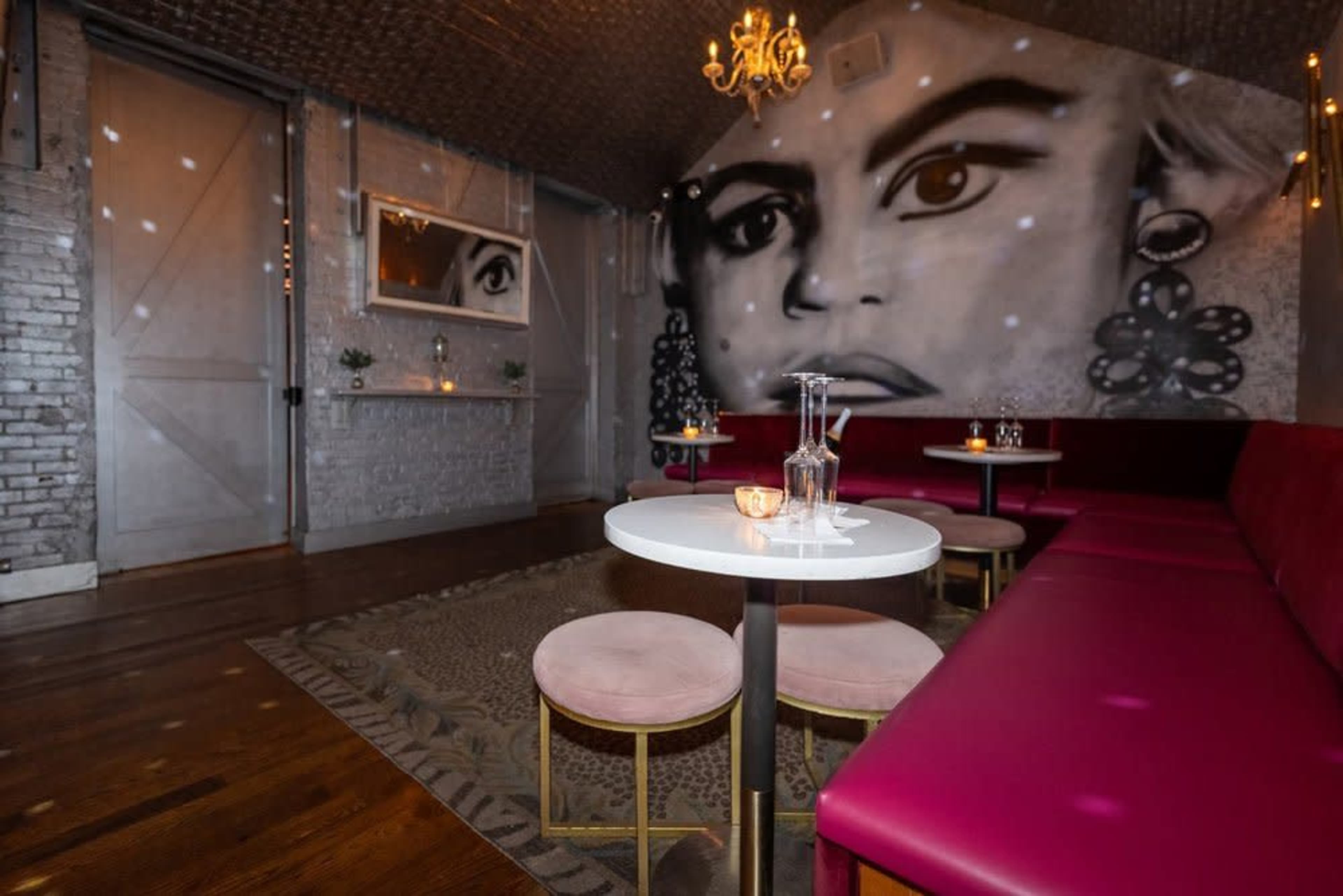 The image shows a stylish lounge with pink seating, a chandelier, and a large mural of a woman's face on the wall.