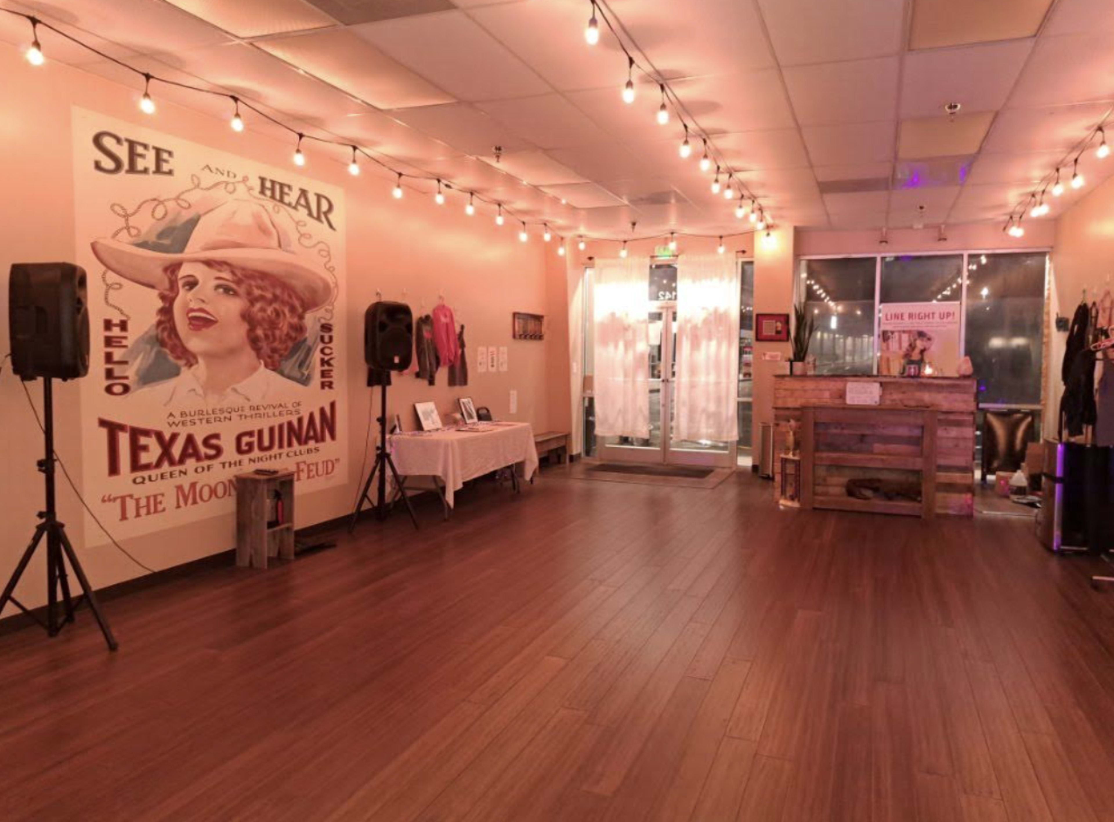 An indoor space featuring warm wooden flooring, a large vintage poster of Texas Guinan on the wall, and string lights illuminating the area.