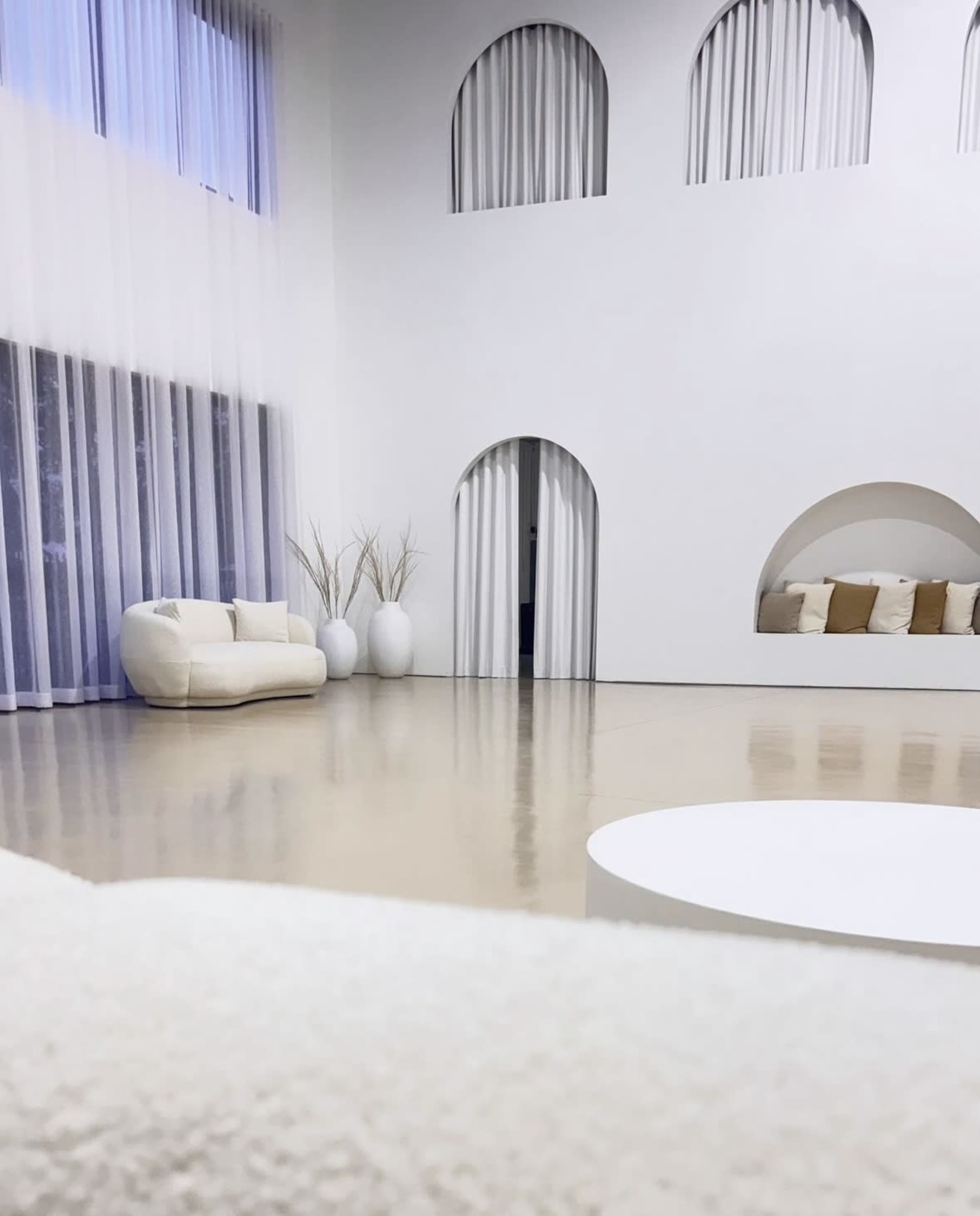 The image shows a spacious, modern room with high white walls, large windows covered by sheer curtains, and minimalist furniture including a light-colored sofa and circular table.